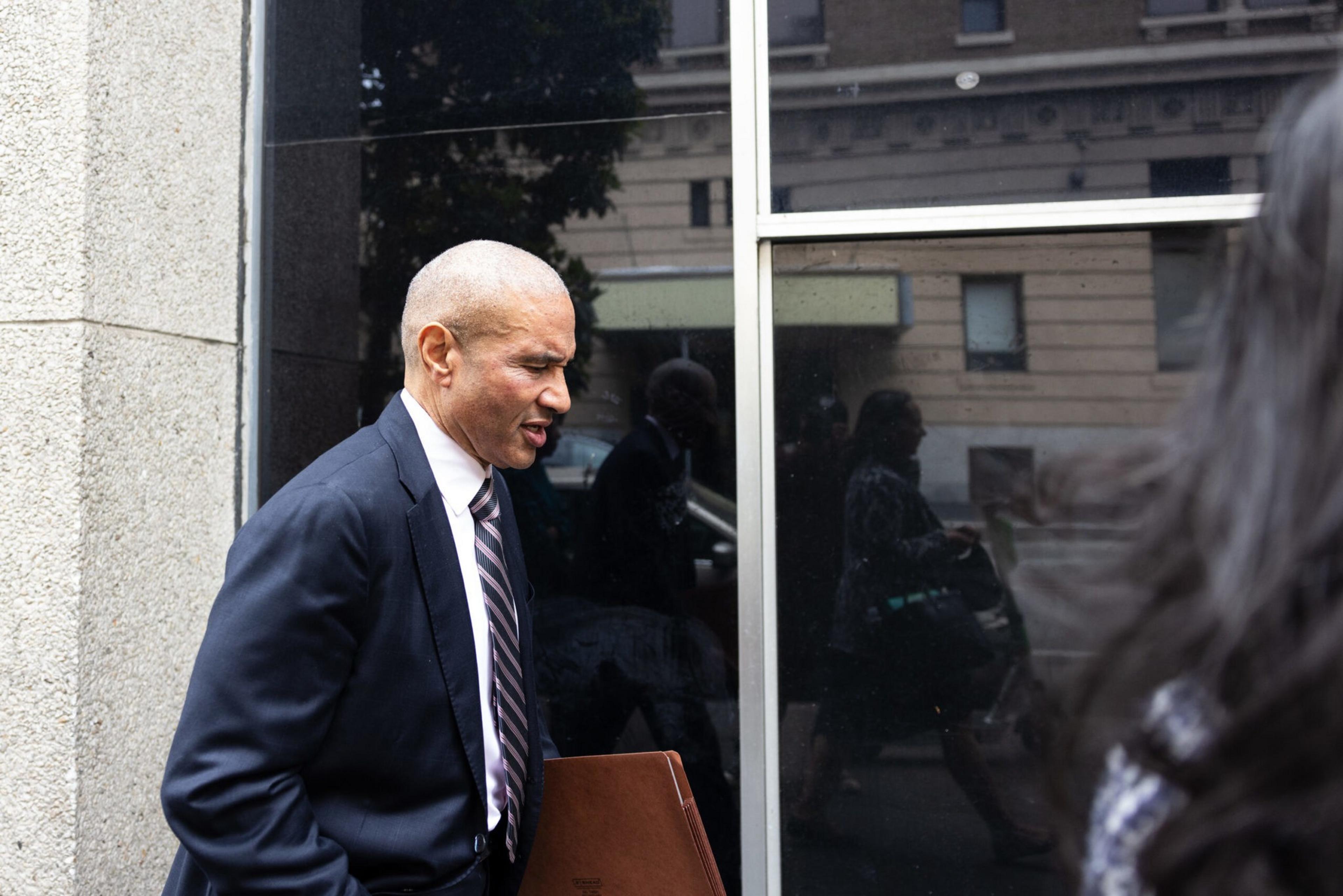 A man in a dark suit with a folder walks by a reflective window.