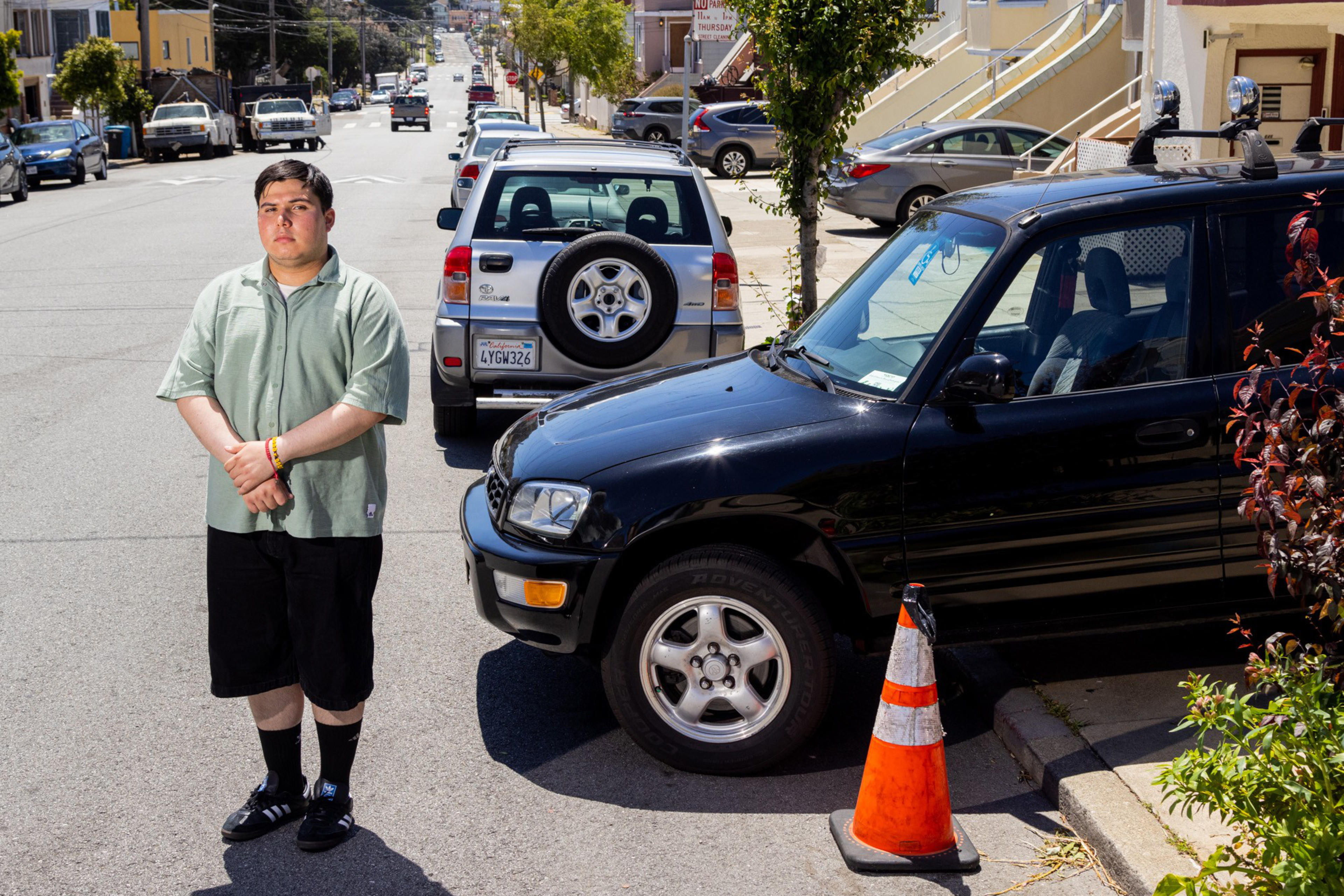 A man stands on a sunny street with parked cars, colorful houses, hills in the background, and a traffic cone nearby.