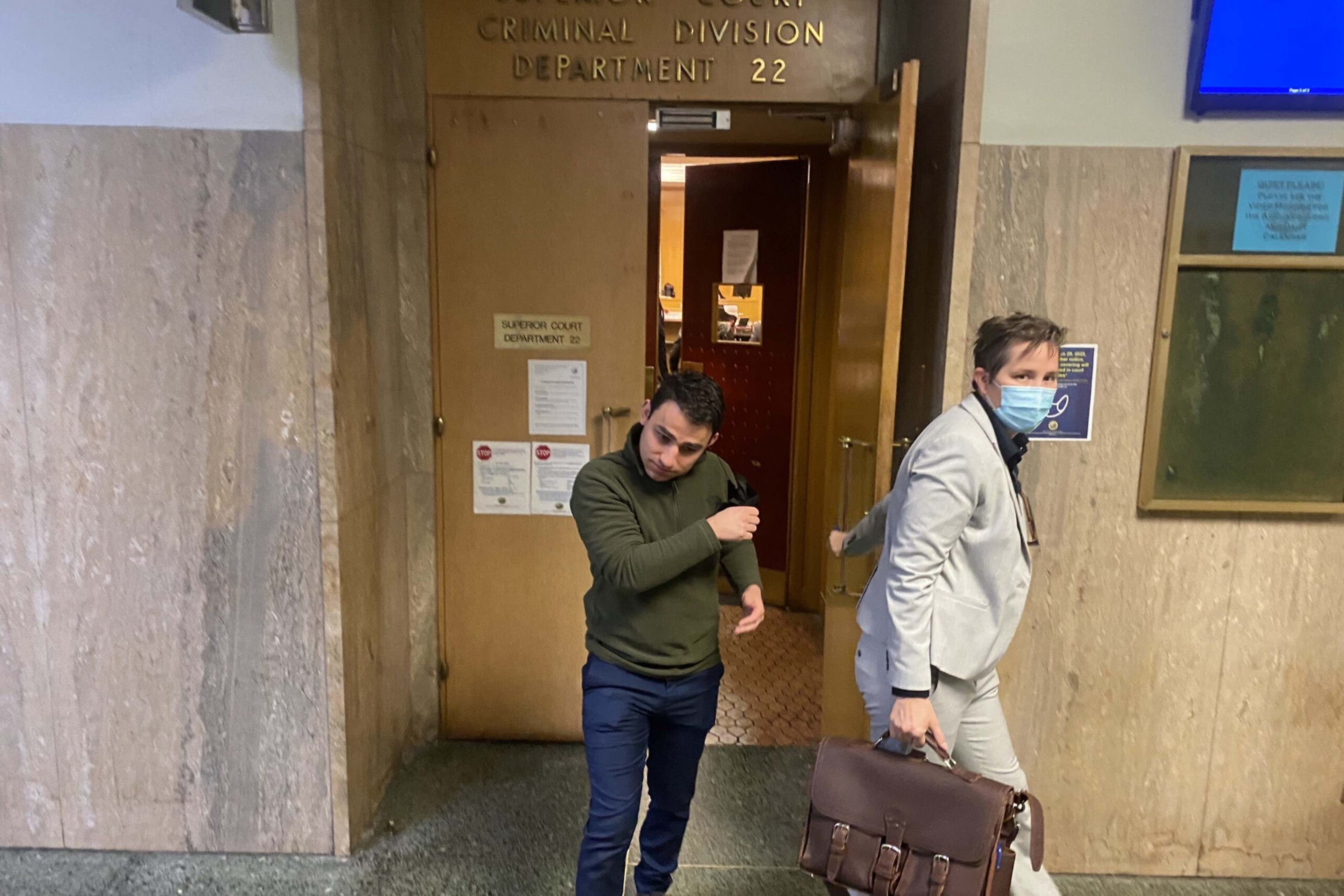 A man in a green sweater and blue pants exits a courtroom door while another man in a light gray suit and face mask carries a briefcase.
