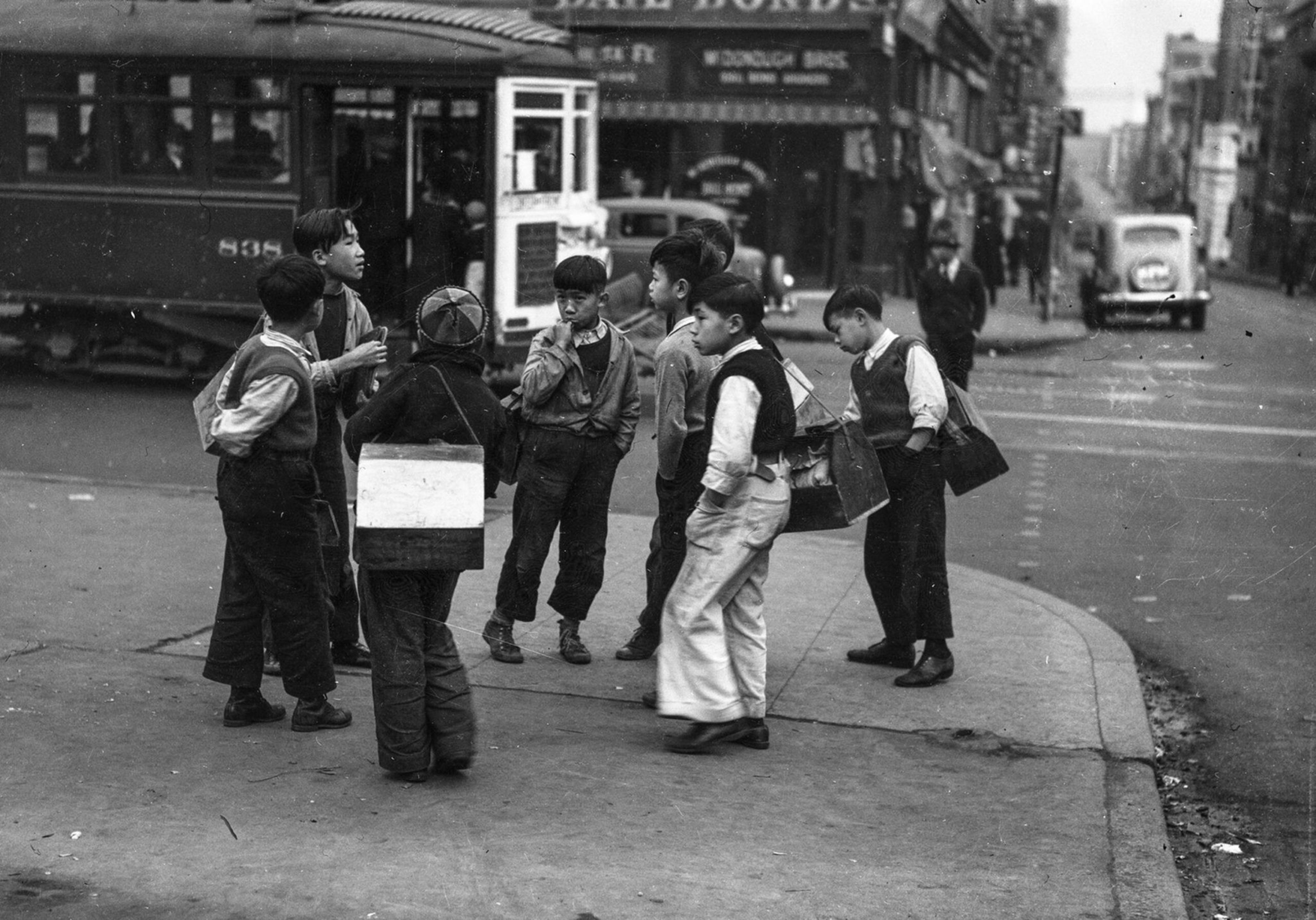 A group of boys, some carrying newspaper bundles, stand talking on a city street corner near a streetcar and vintage cars.