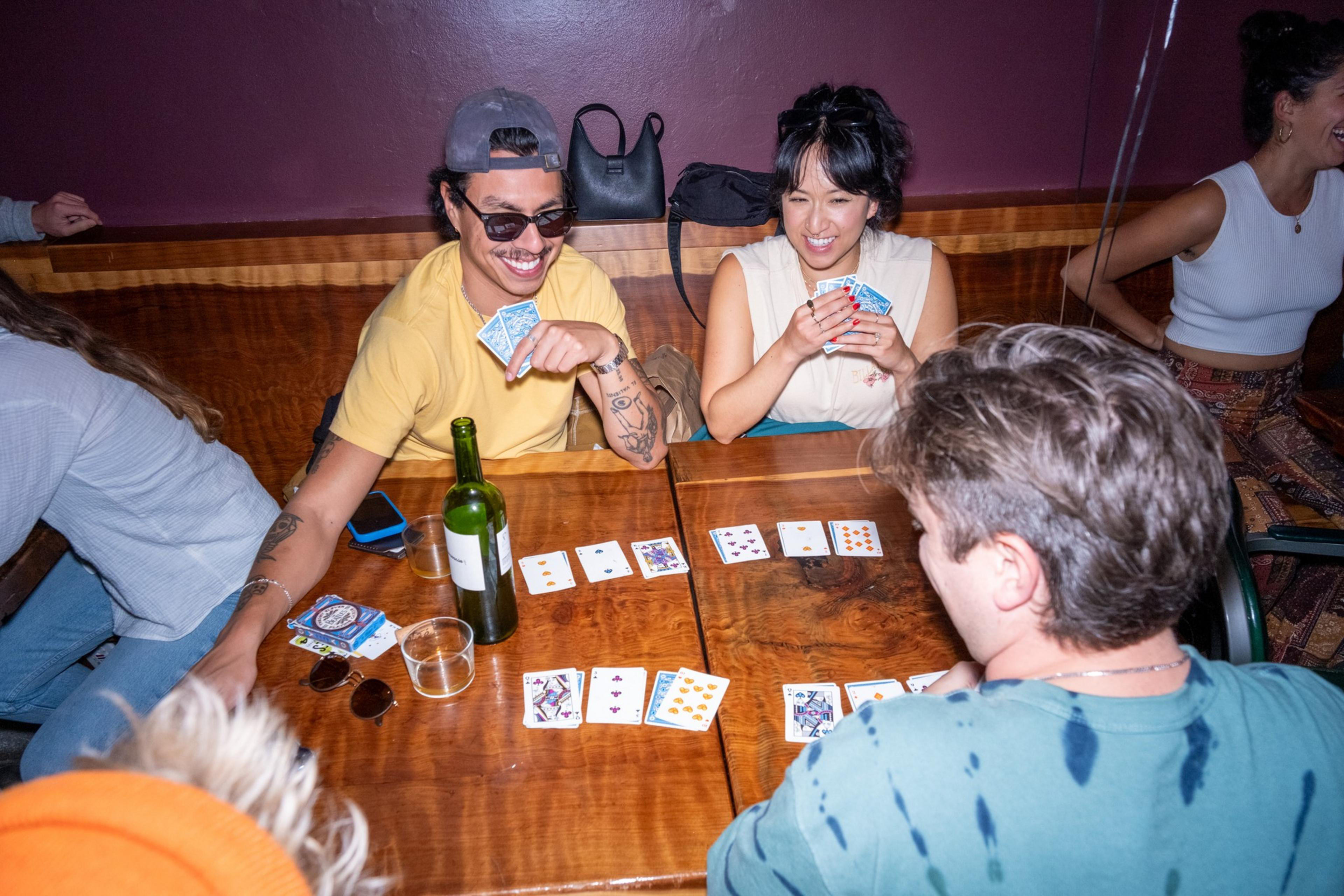 People are playing cards at a wooden table, laughing and enjoying themselves. There's a bottle and some drinks on the table, and the atmosphere is relaxed and fun.