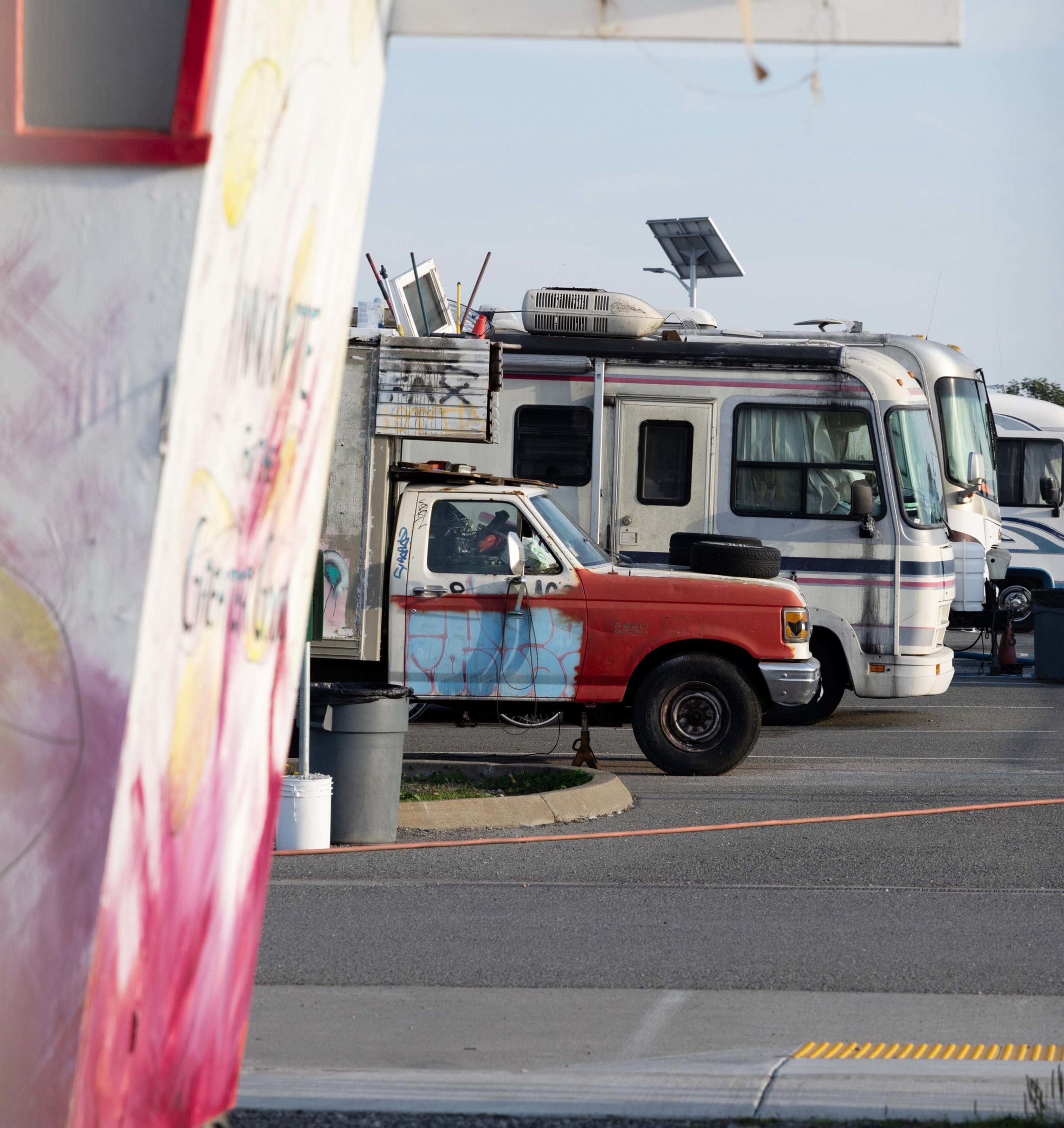 A parking lot with a red, graffiti-covered truck and several RVs parked beside it. A painted wall is partially visible on the left.