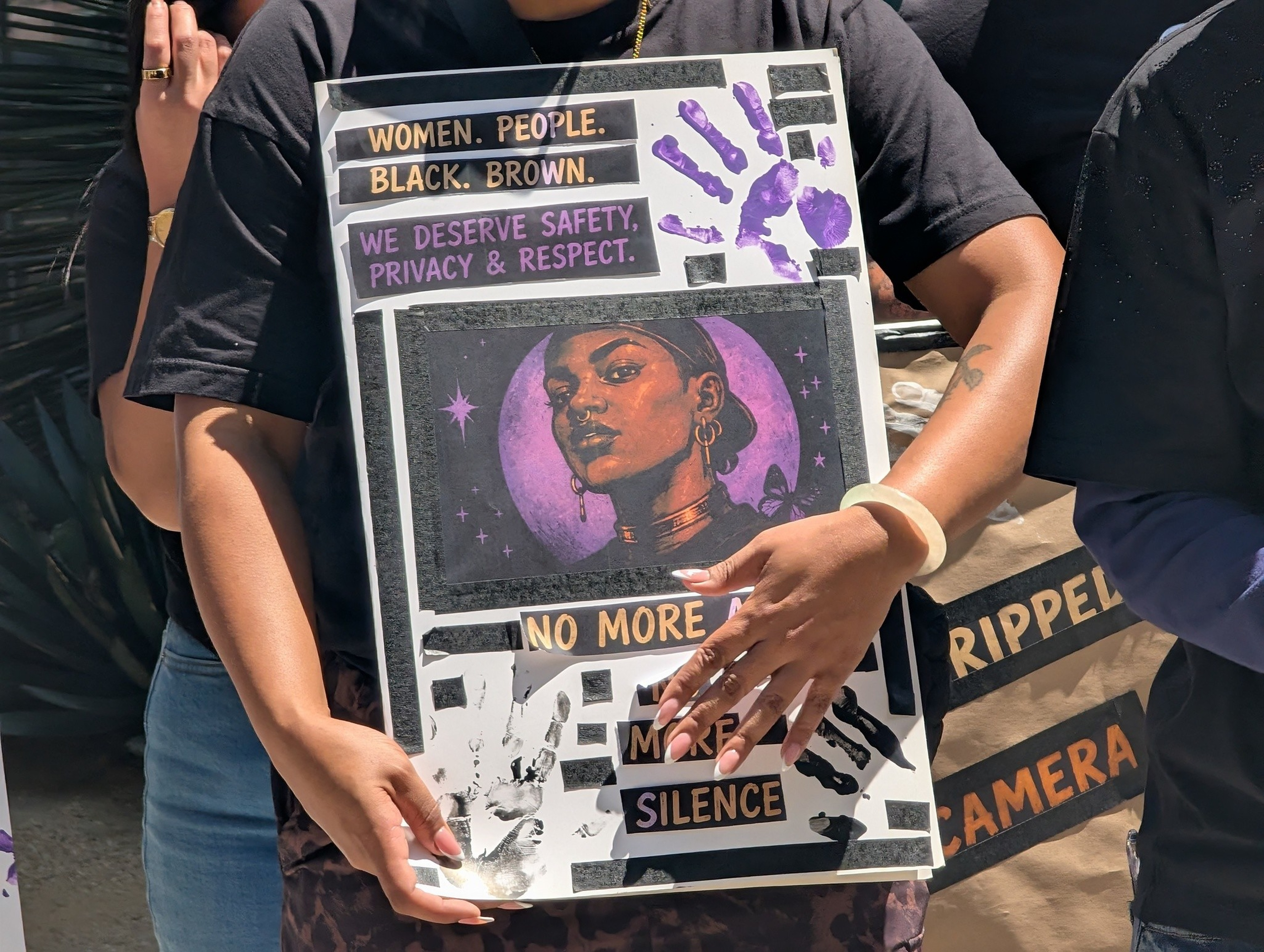 A person holds a sign with a purple-toned portrait of a Black woman, featuring phrases like “Women. People. Black. Brown.” and “No more silence.”