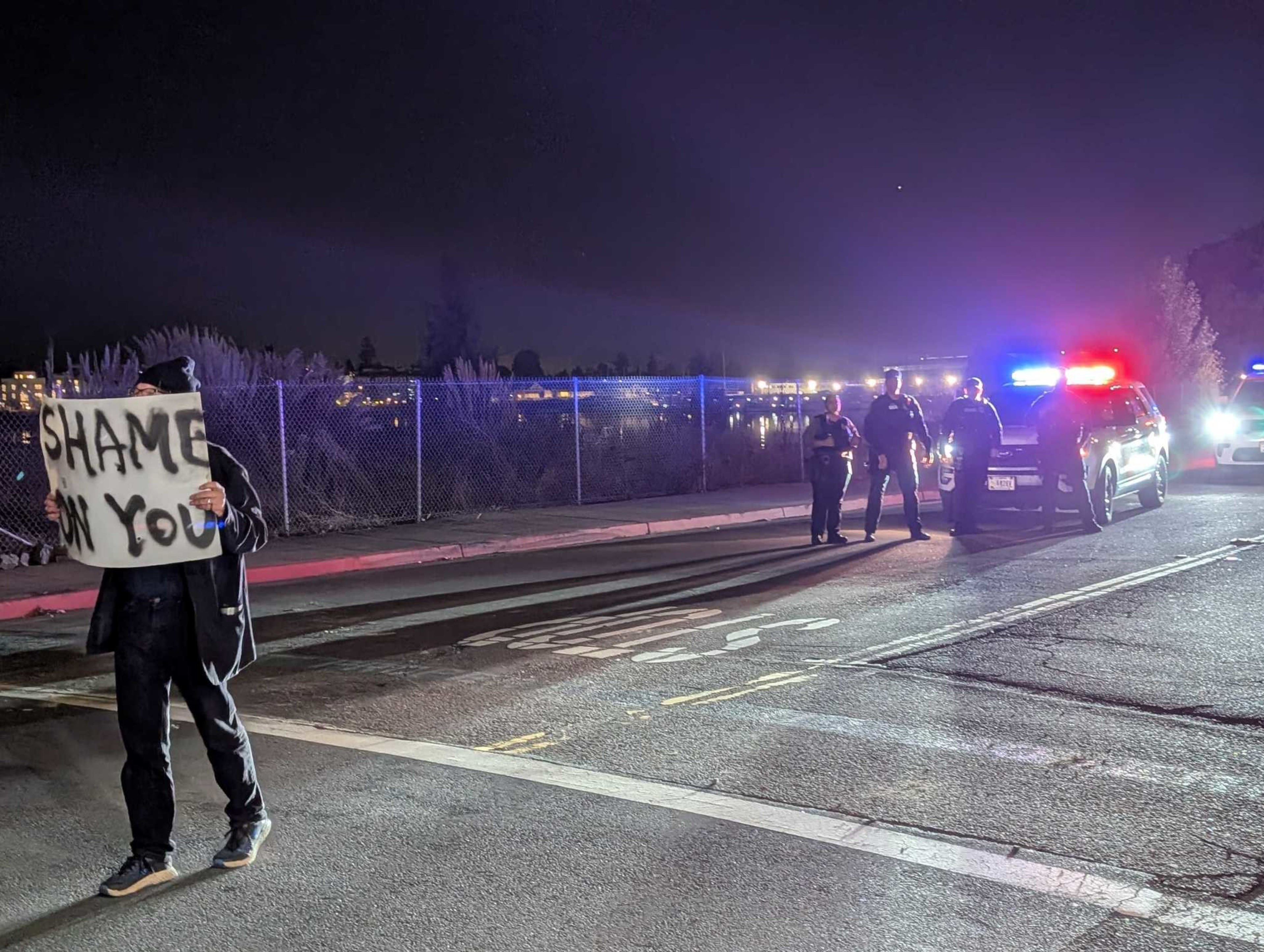 A person holds a "SHAME ON YOU" sign on a street at night while three police officers stand near a police car with flashing lights.