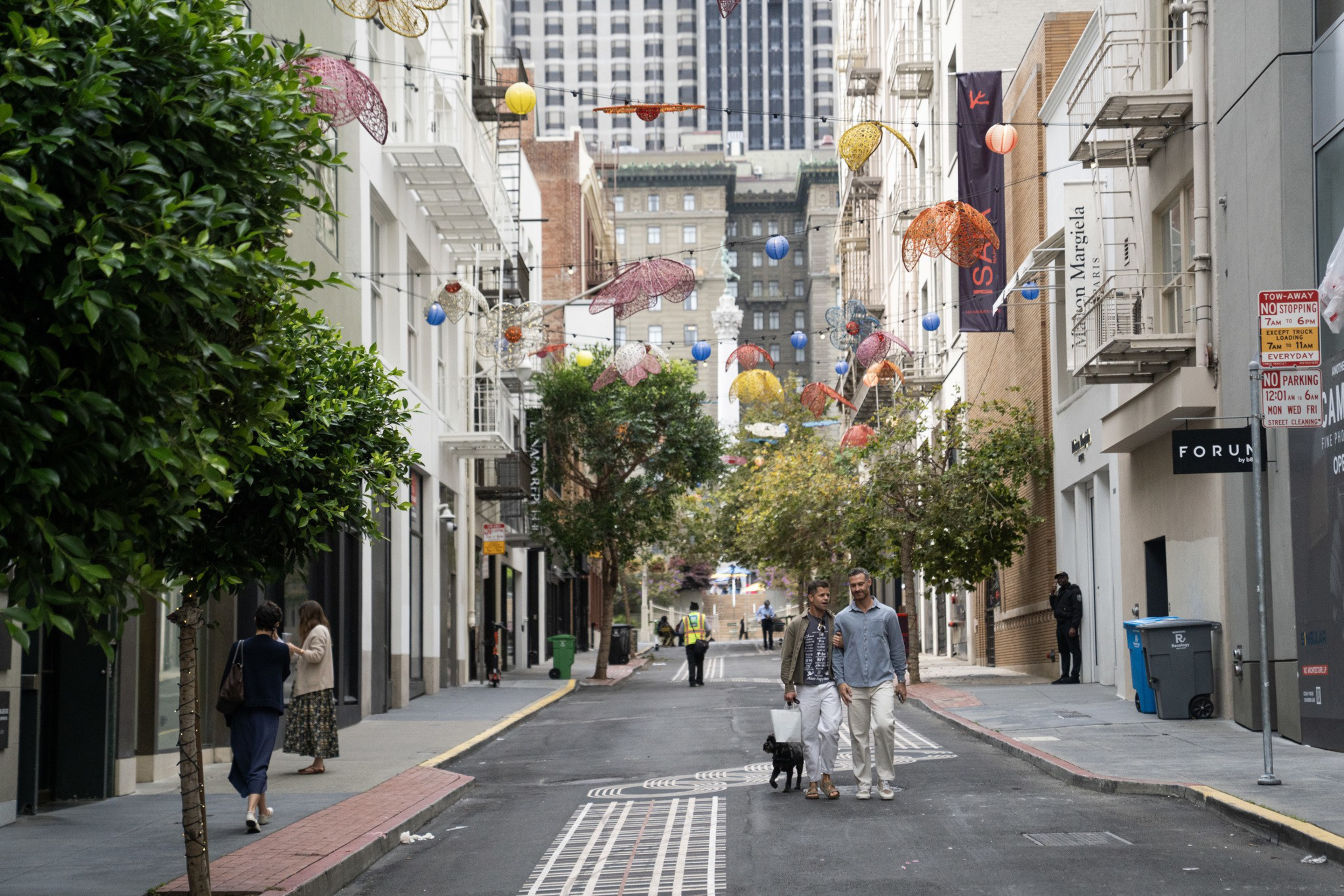 A city street with trees and colorful umbrella decorations hanging overhead shows people walking, including a couple with a small black dog.