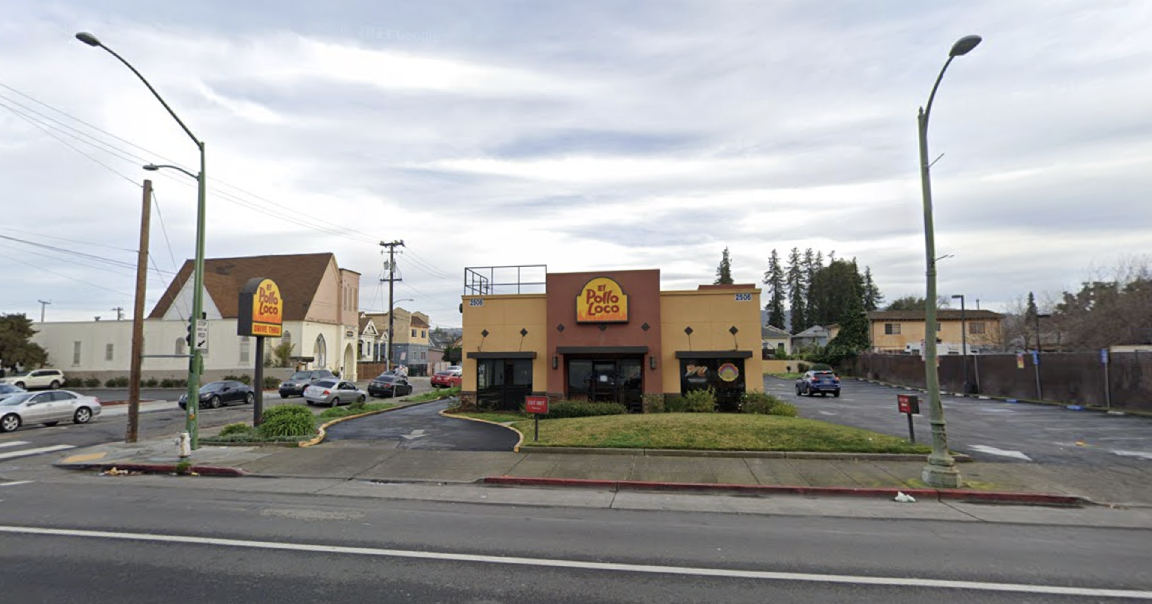 A small El Pollo Loco restaurant with a yellow and orange exterior sits between two parking areas, under a cloudy sky with street lamps nearby.