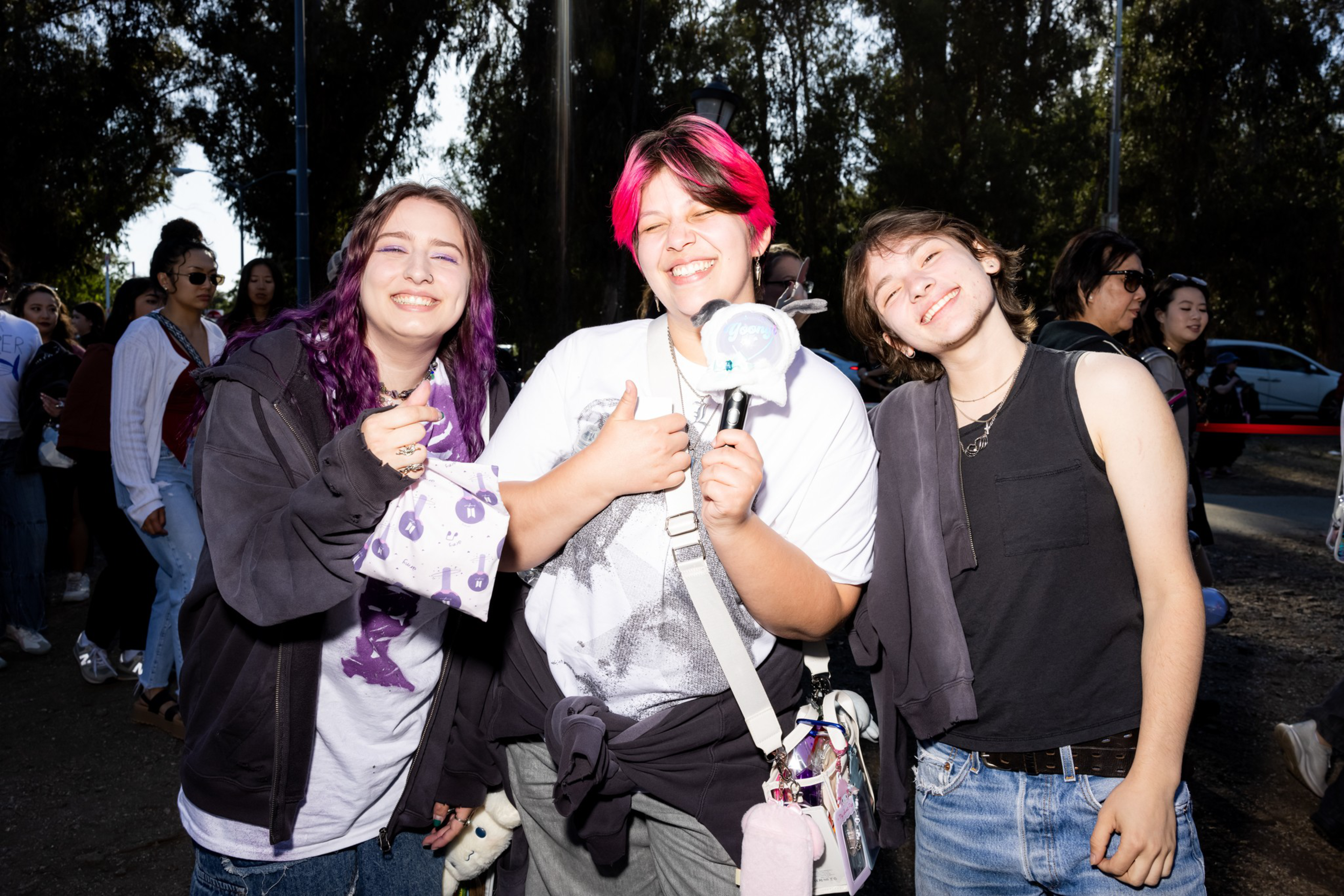 Three smiling friends stand close together outdoors, one with bright pink hair holding a light stick, surrounded by other people in casual attire.