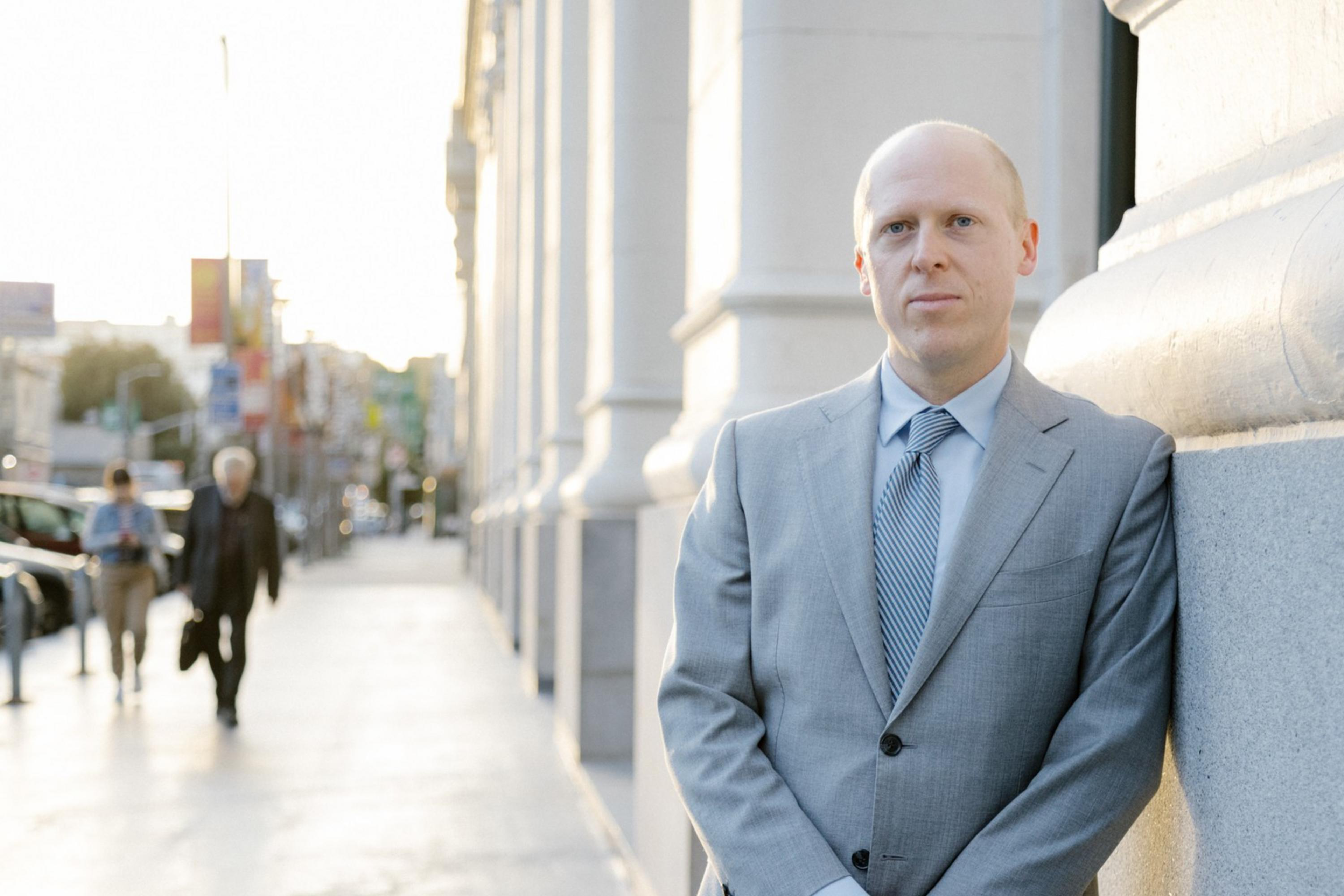 A man in a gray suit stands by a street, leaning against a building, with pedestrians in the soft-focus background.
