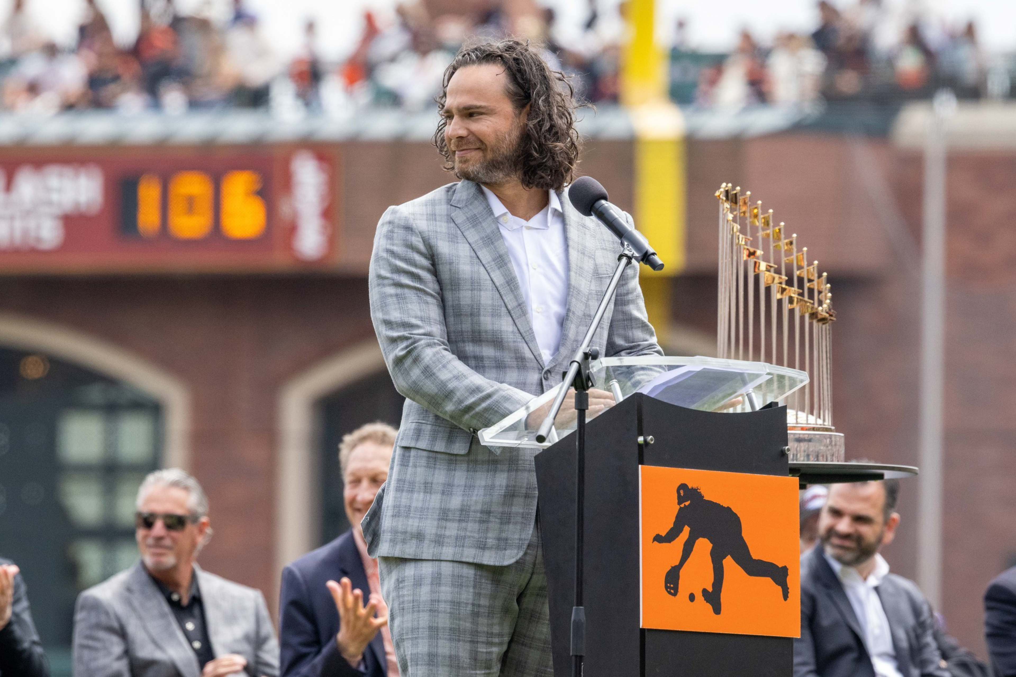 A man in a gray plaid suit stands at a podium with a microphone and a trophy beside him. People clap in the background, and thereâ€™s a logo on the podium.