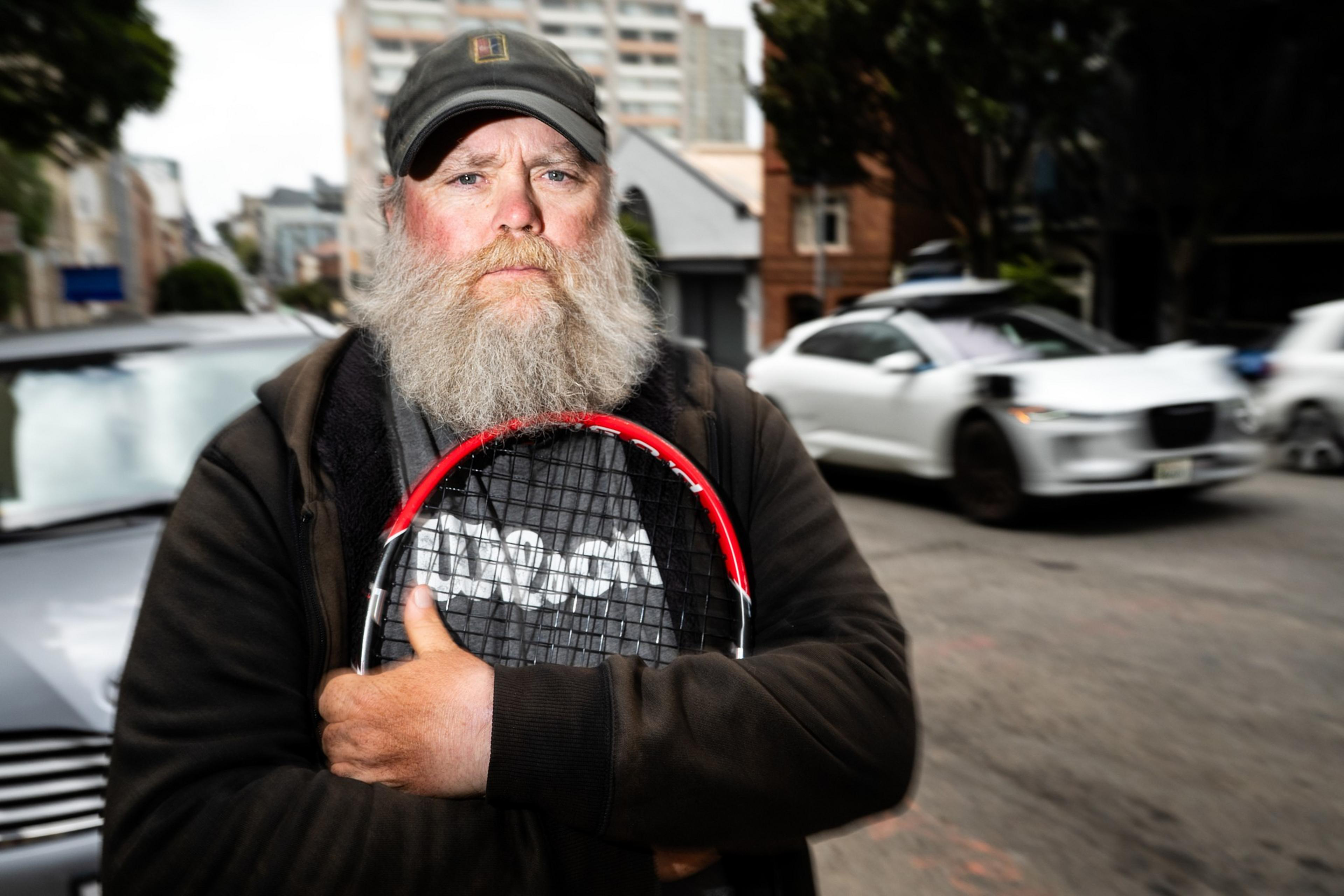 A bearded man in a cap holds a tennis racquet on a street, with blurred cars and buildings in the background, creating a dynamic city scene.