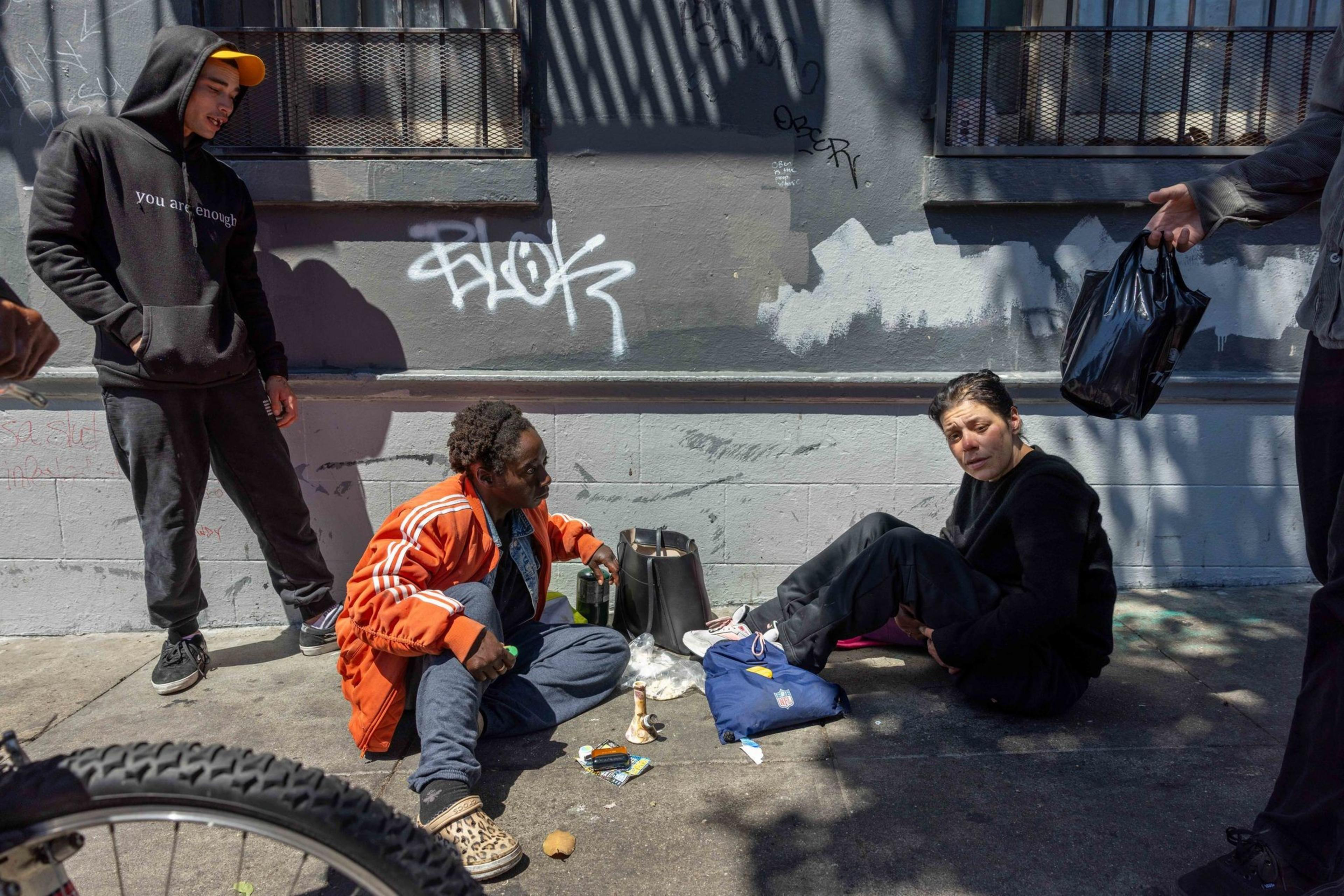 Three people are on a graffiti-marked sidewalk. Two sit on the ground with belongings, while one stands nearby. Another hand extends holding a black bag.