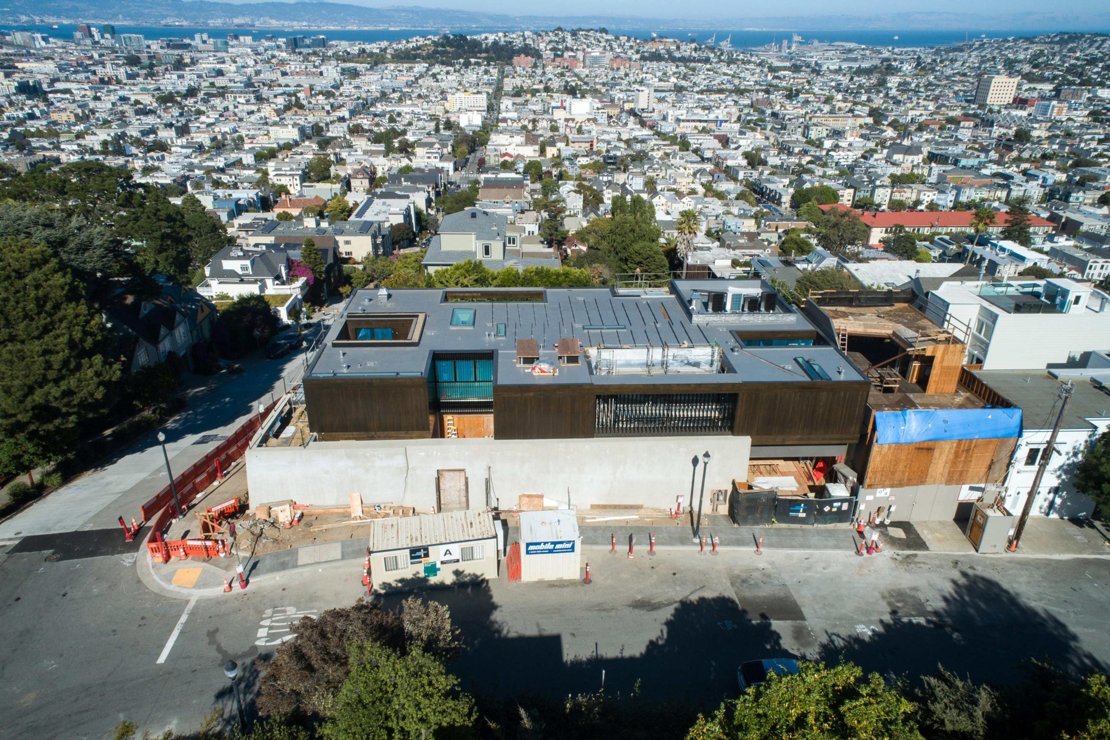 An aerial view of a cityscape, focusing on a large modern house under construction with surrounding residential areas and distant water in the background.