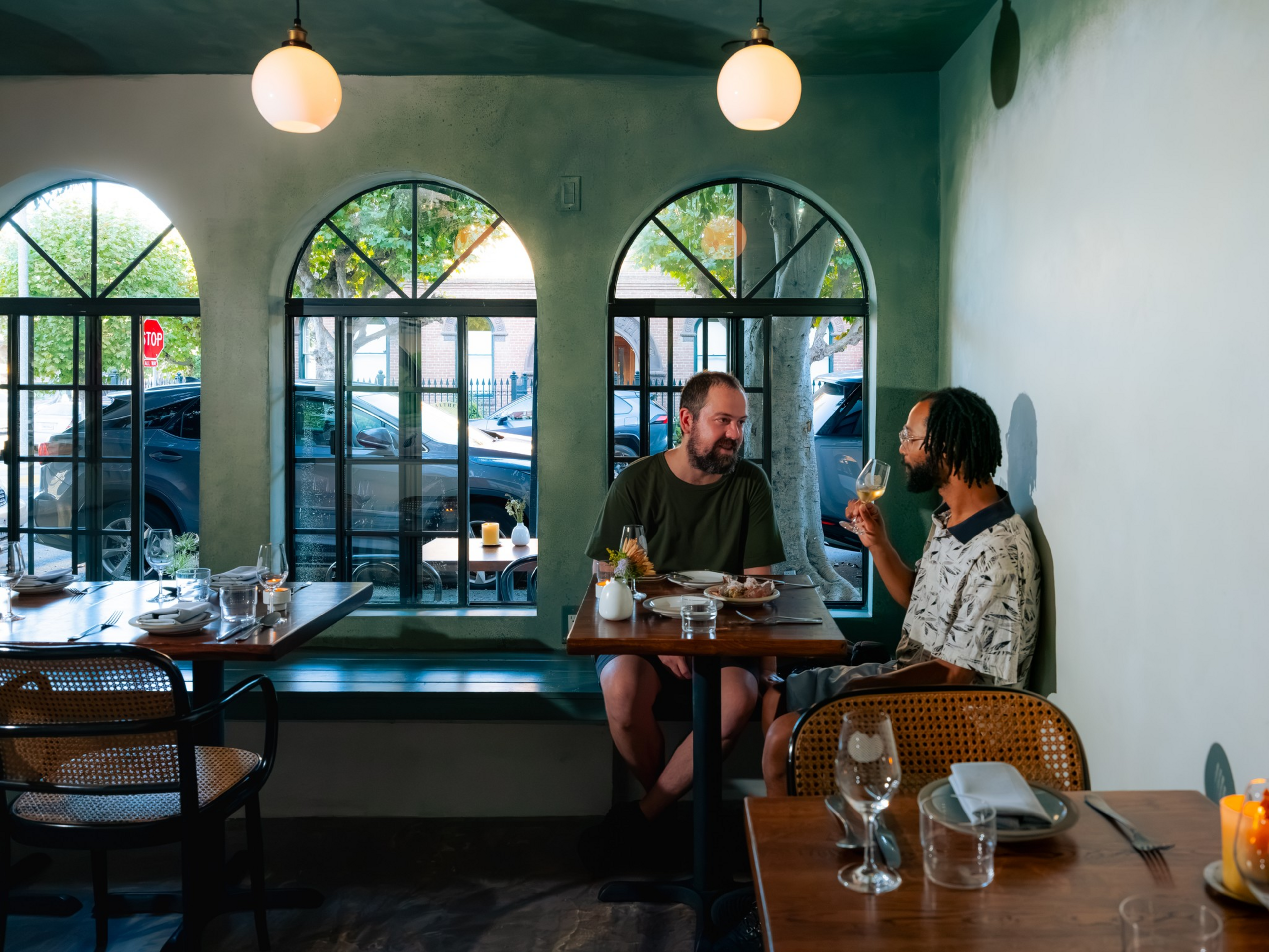Two men sit at a small wooden table by arched windows in a cozy ...