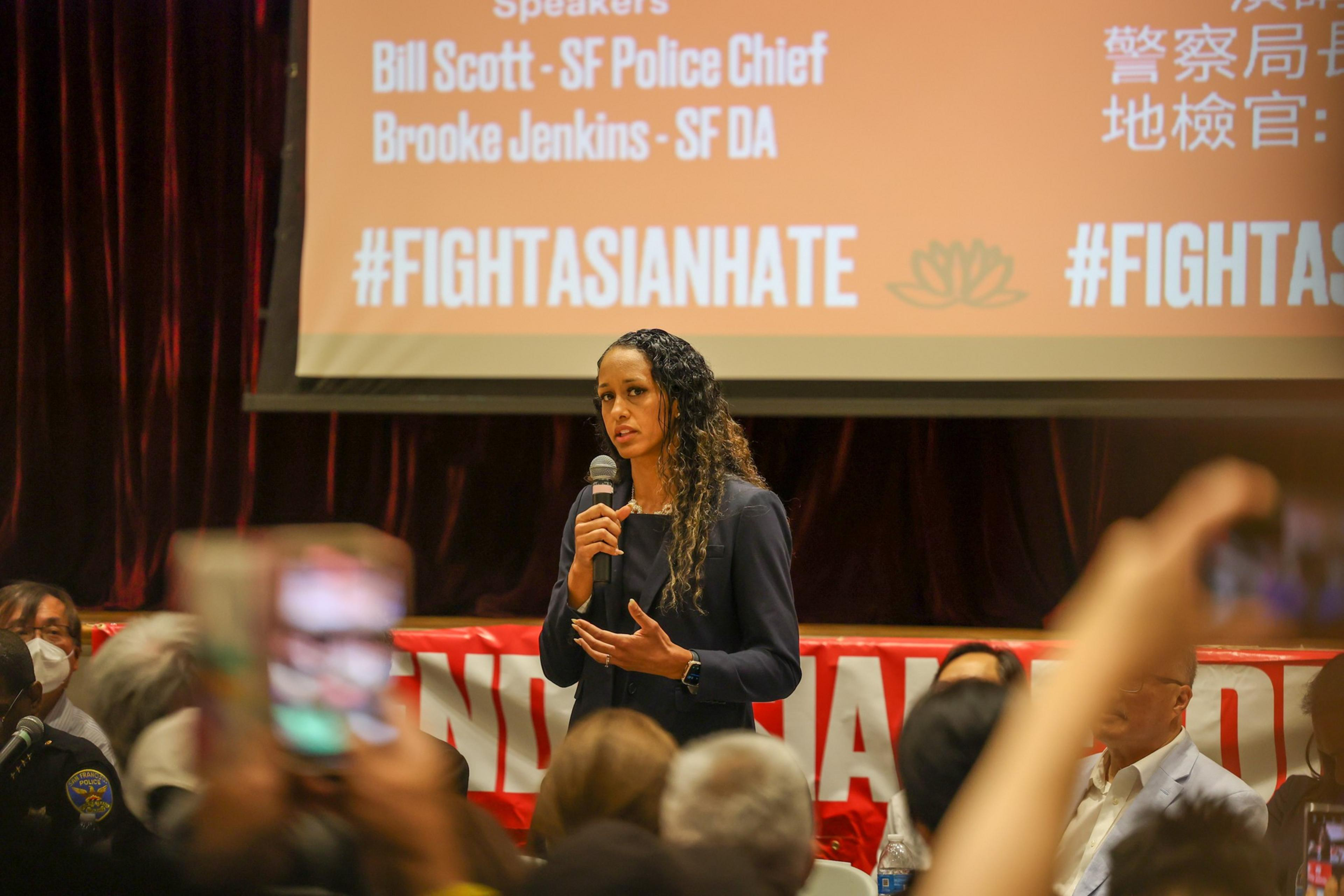 A woman speaks at a podium, with a backdrop reading "#FIGHTASIANHATE", as an audience with phones captures the moment.