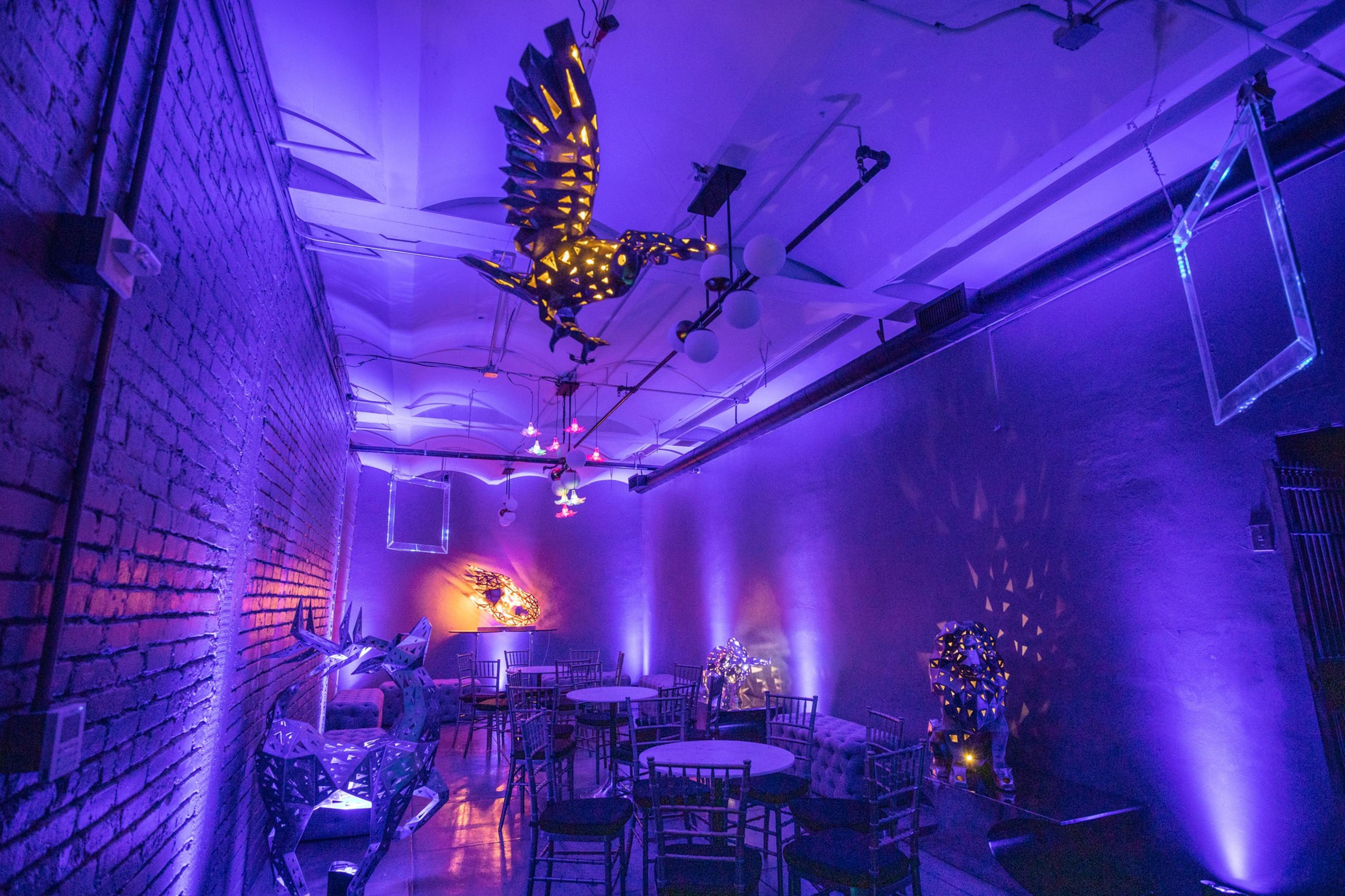 The room features purple-lit walls, modern art sculptures, round tables with chairs, a brick wall, and a ceiling adorned with an illuminated bird figure.