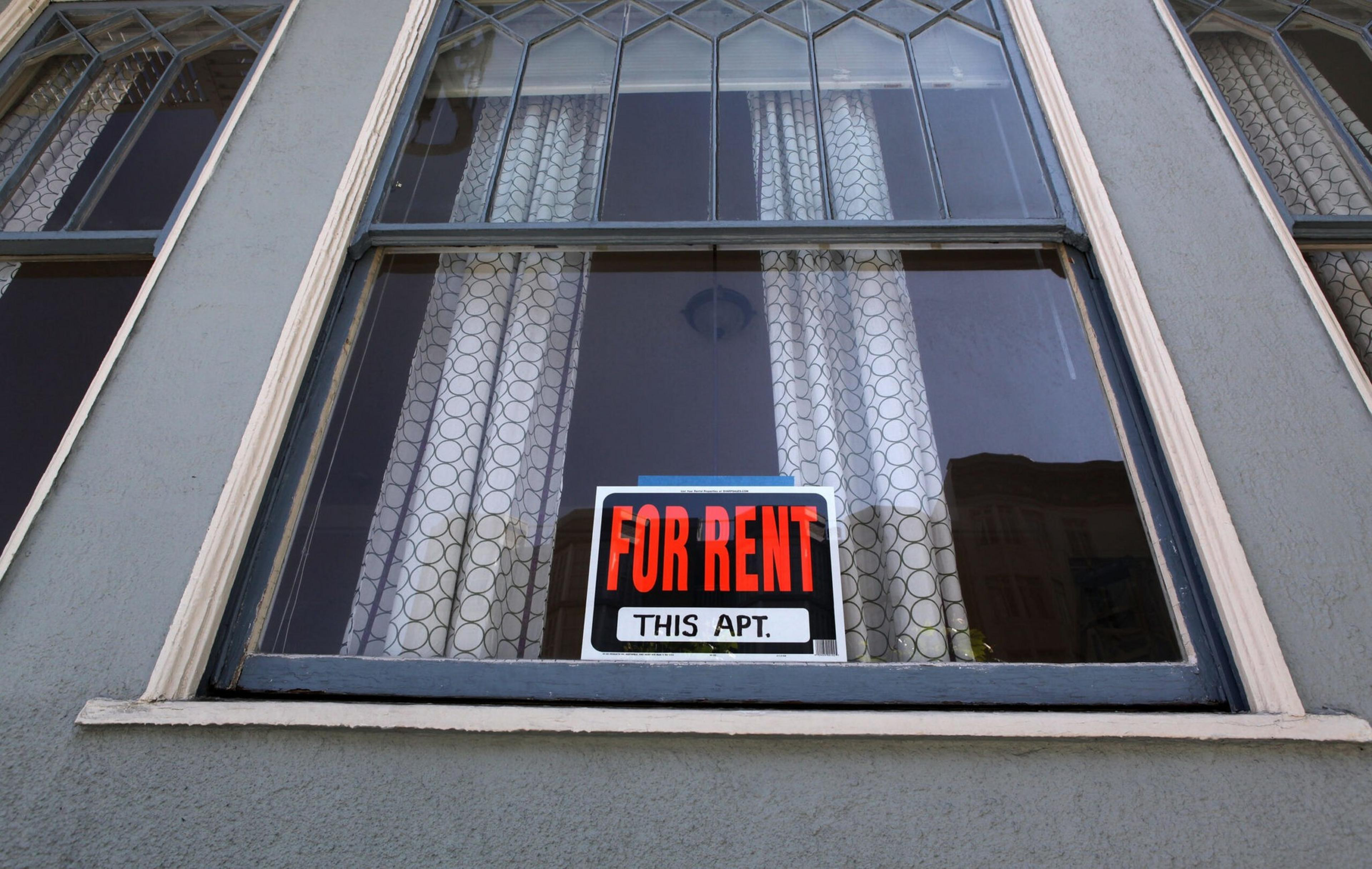 A window with white curtains behind glass has a sign that reads "FOR RENT THIS APT." in bold red and black letters.