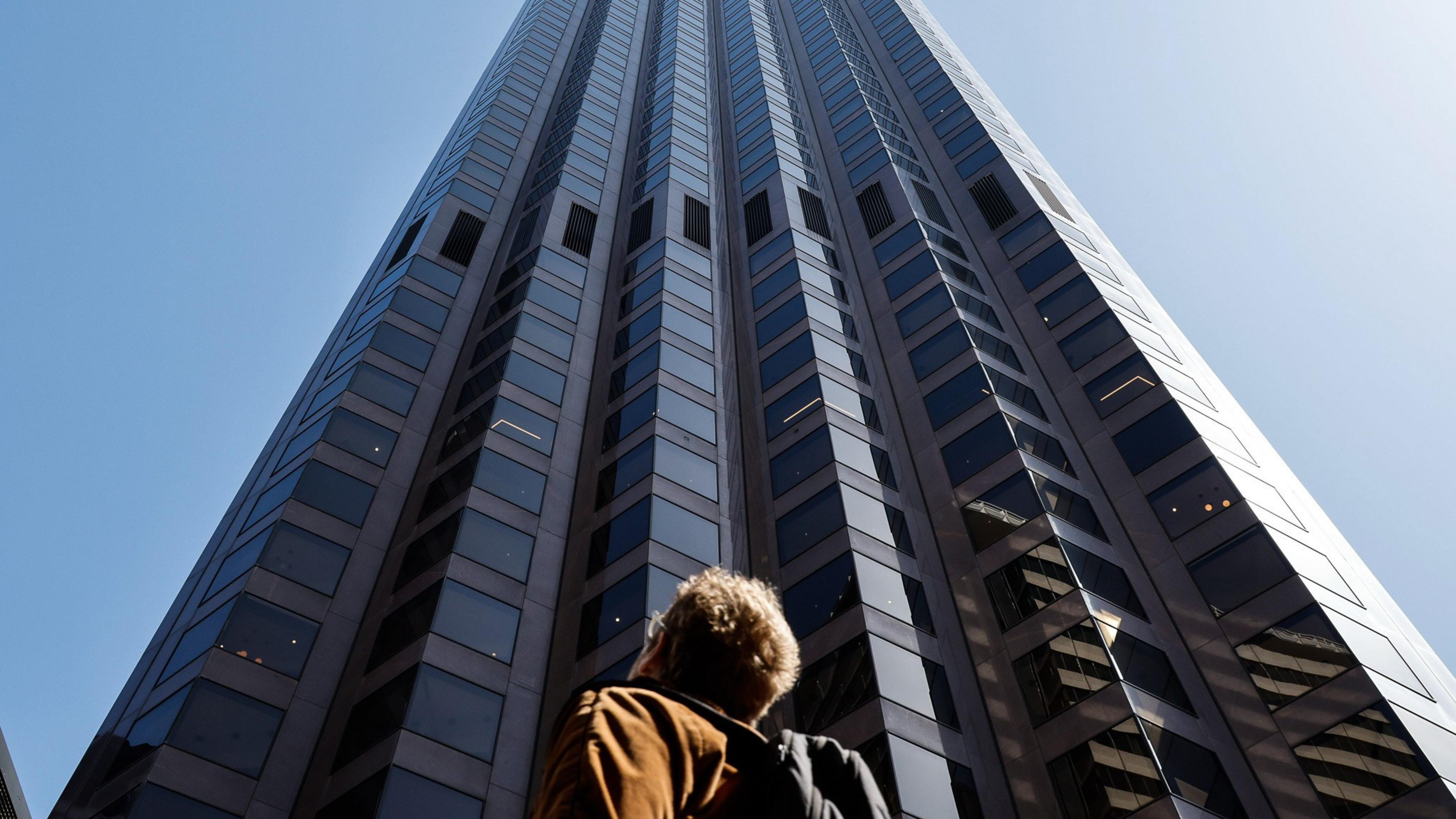 A person looks up at a towering skyscraper with mirrored windows, reflecting the clear blue sky above.