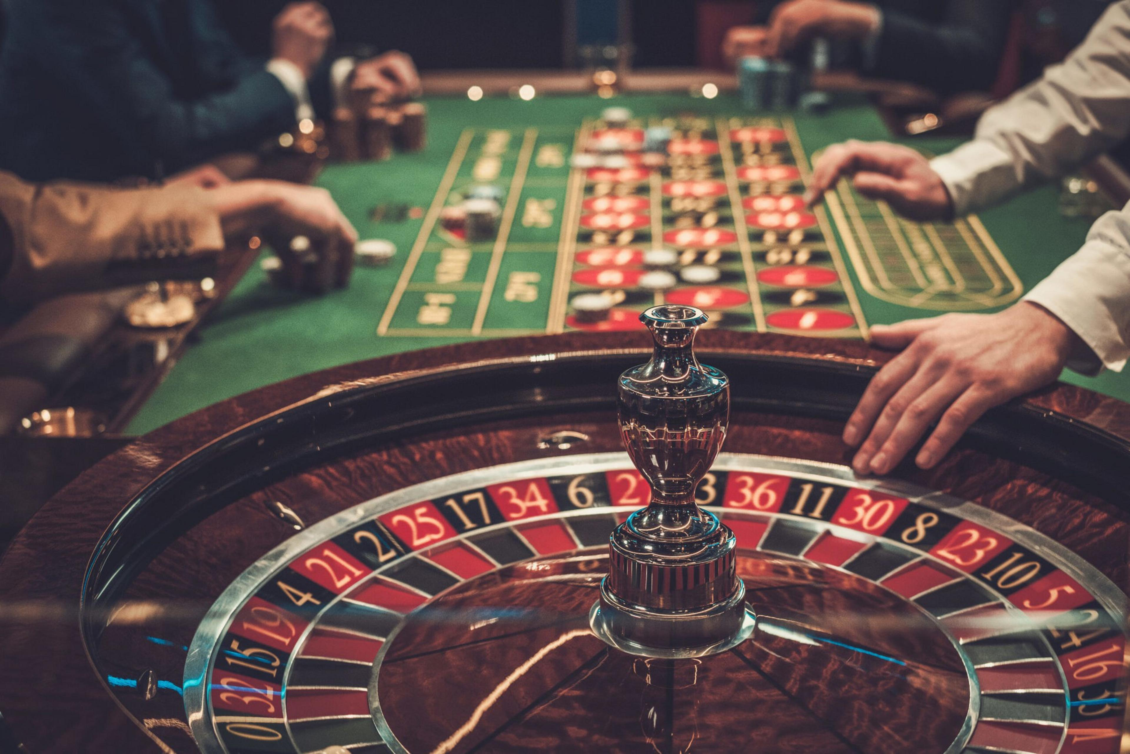A roulette wheel spins in the foreground while several players place bets and chips on a green betting table in the background.