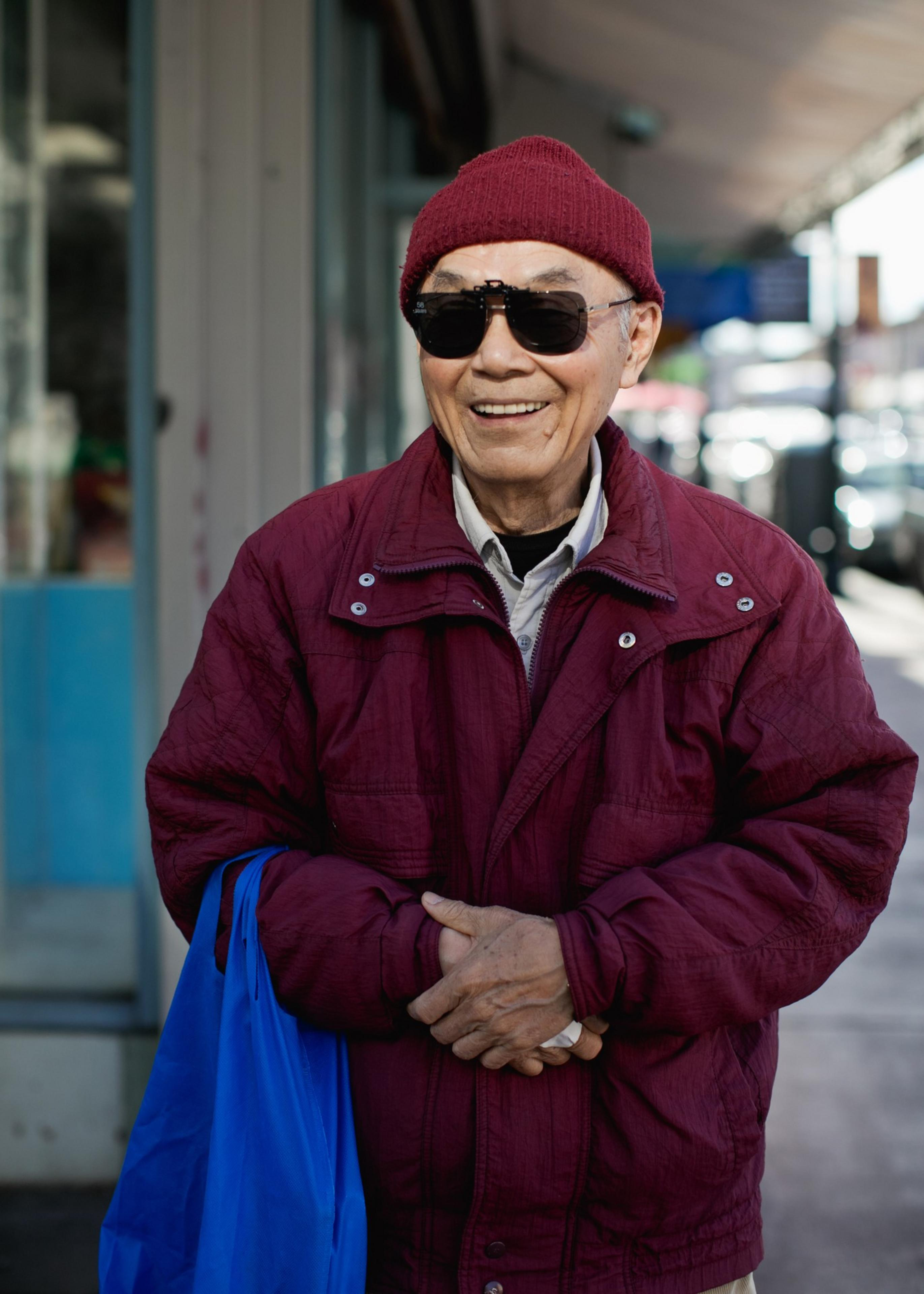 An elderly man wearing a burgundy jacket and beanie, dark sunglasses, smiles while holding a blue reusable shopping bag on a city sidewalk.