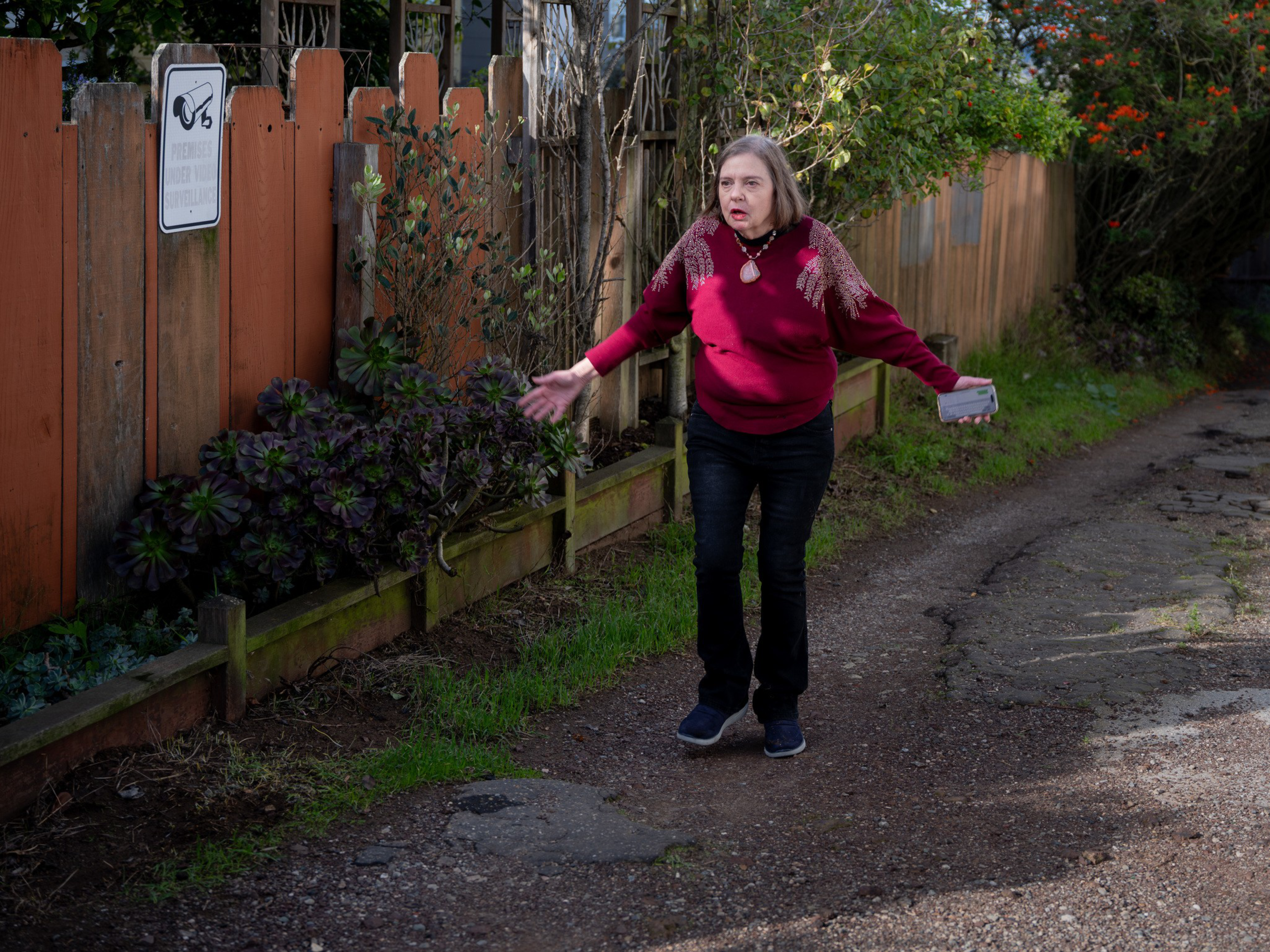 A woman in a red sweater stands on a dirt path beside a wooden fence, holding an object and gesturing with an expressive face.