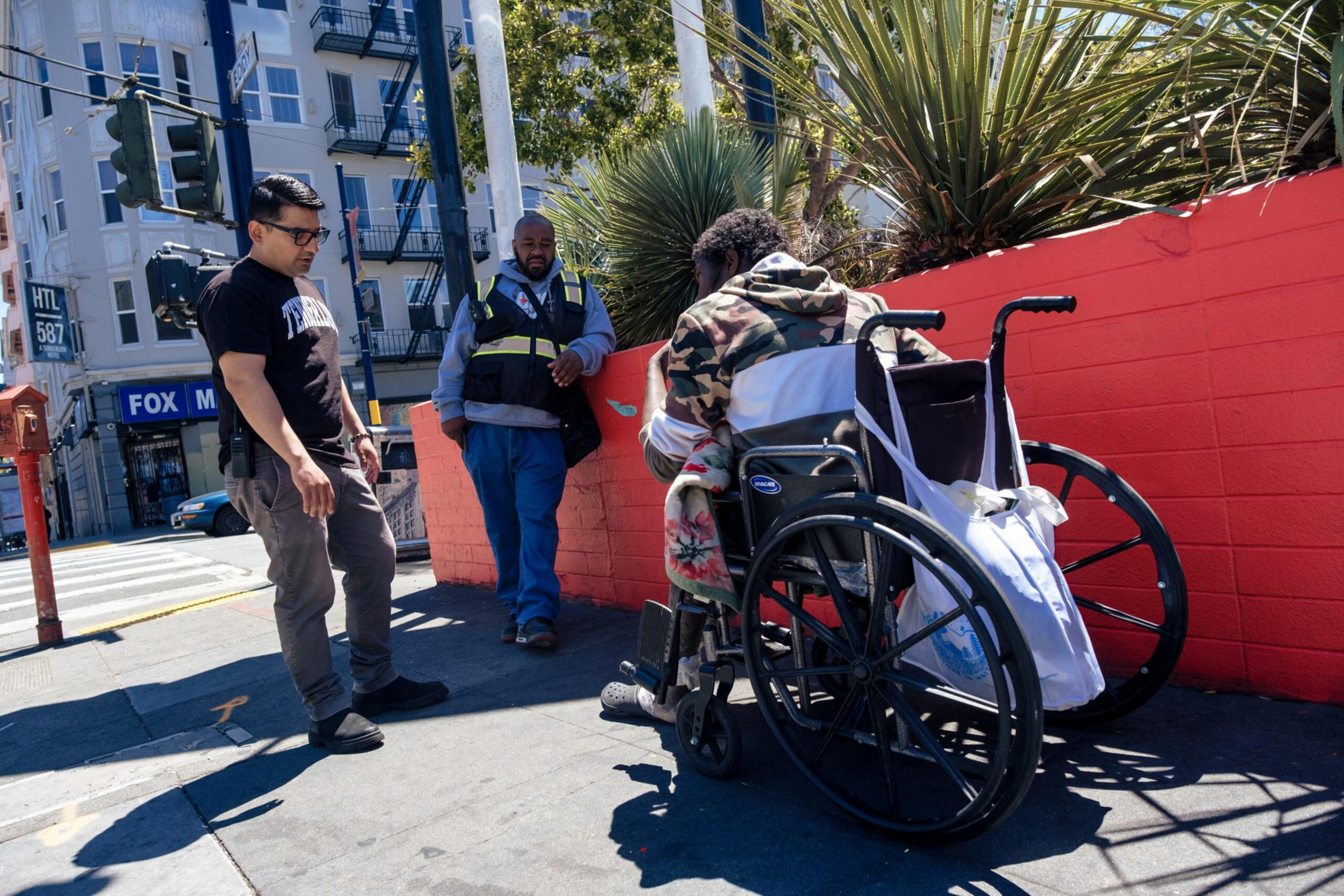 A person in a wheelchair with a camouflage jacket faces a red wall while two men, one in casual clothes and one in a safety vest, stand nearby on the sidewalk.