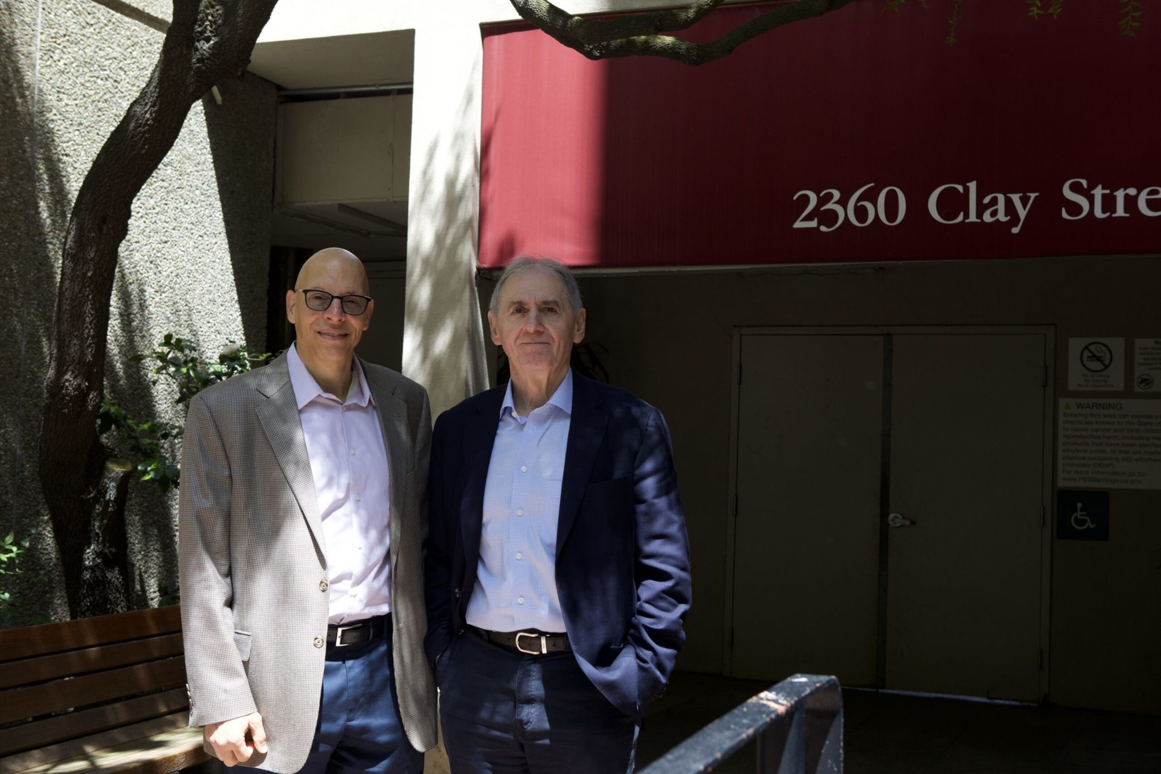 Two men wearing suits stand side by side outside a building with a red awning that reads "2360 Clay Street." One man is bald with glasses, the other has gray hair.