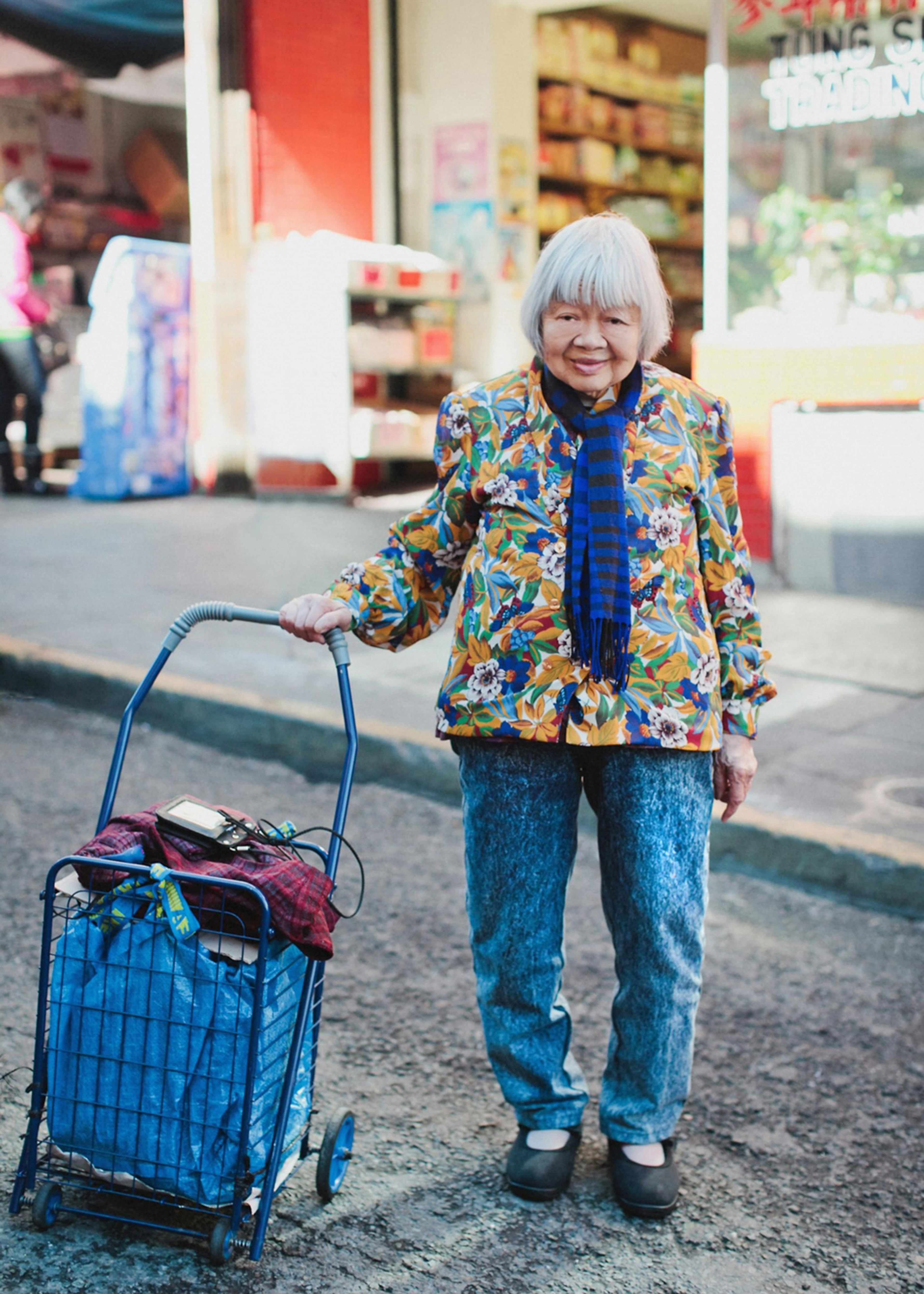 An elderly woman with white hair wears a floral jacket, blue jeans, and a striped scarf, holding a blue shopping cart on a city street.