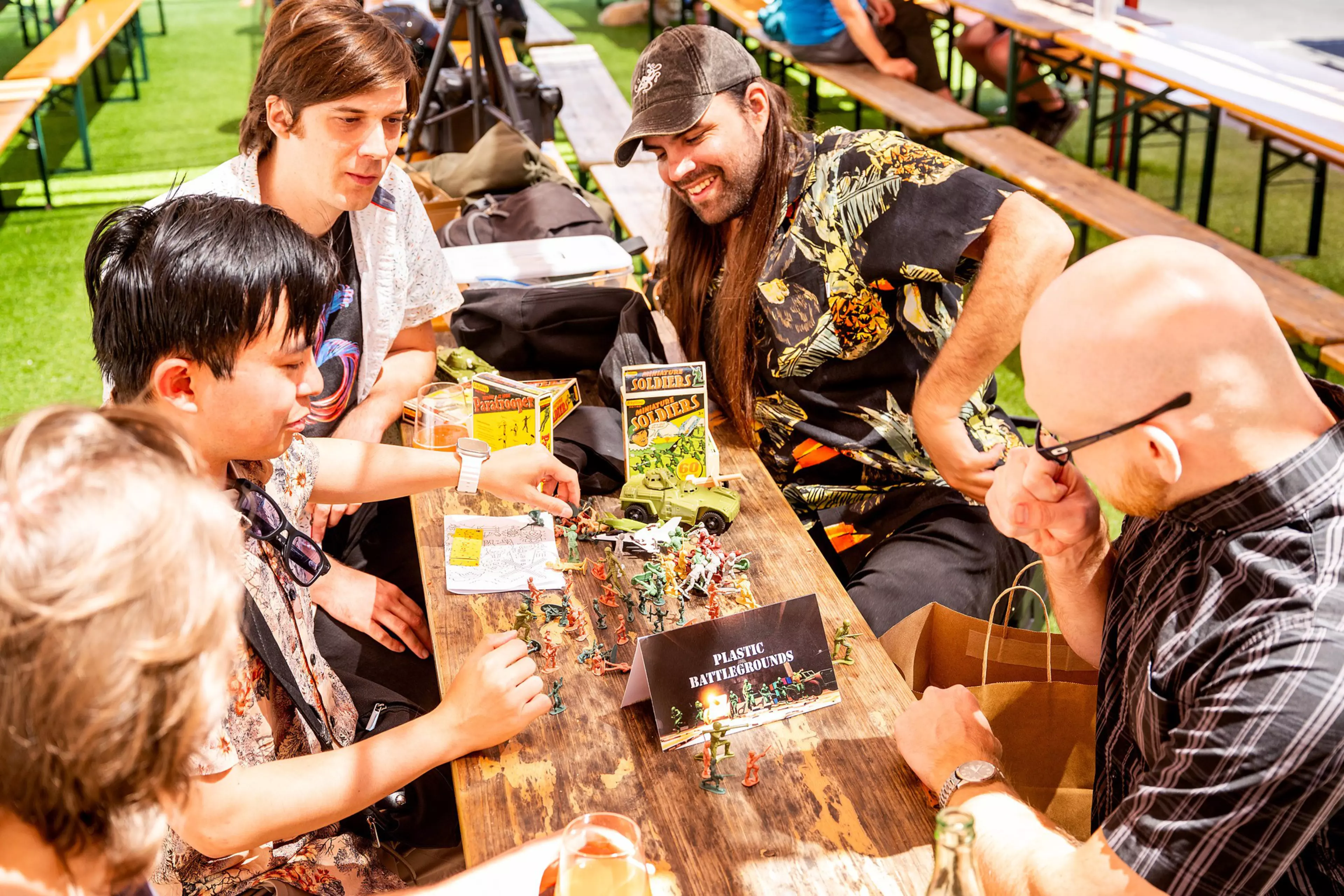 Five people sit around a wooden table outdoors, playing with miniature toy soldiers and vehicles labeled "Plastic Battlegrounds."