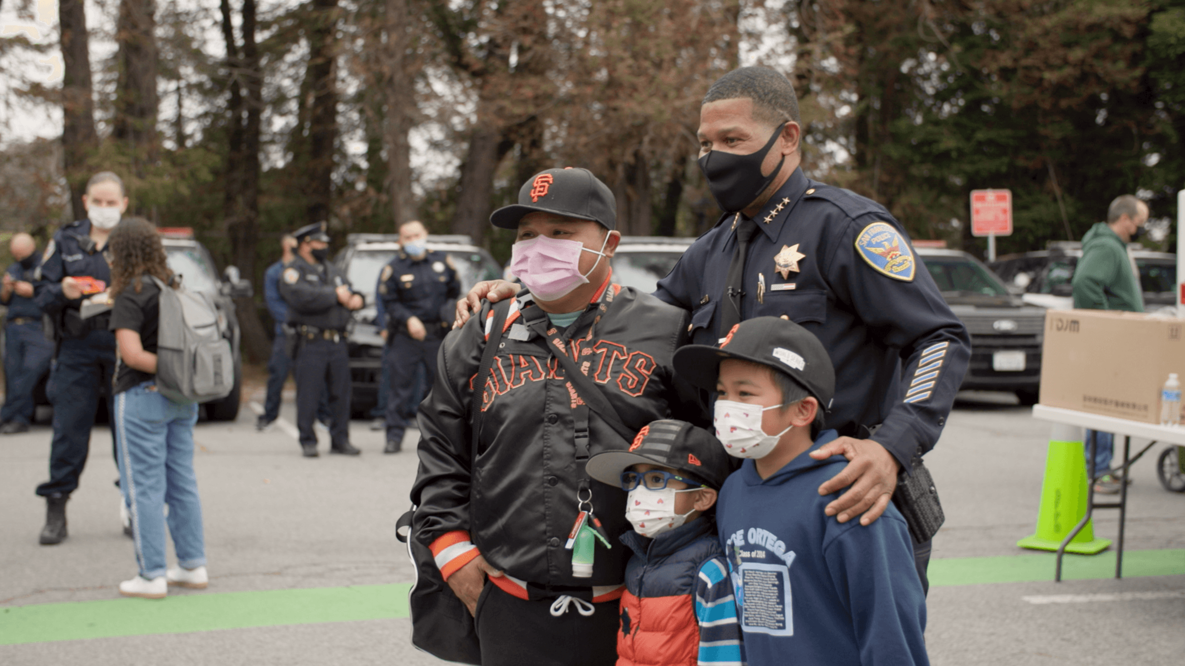 A police officer in uniform poses with a man and two children wearing masks and baseball caps, with several officers and people in the background.