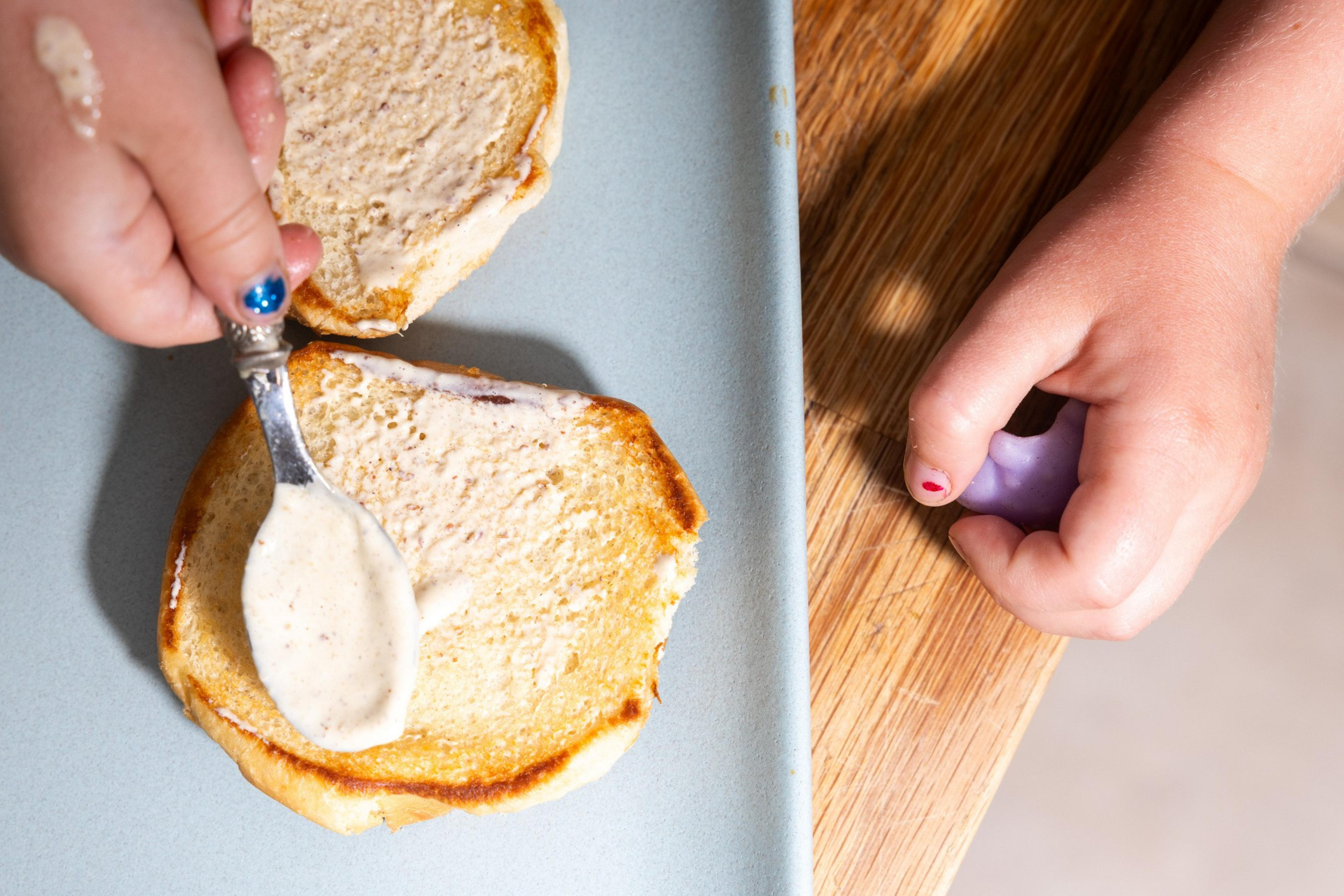 A child spreads creamy sauce on a toasted bun with a spoon. One hand holds the spoon, and the other rests on a wooden table, gripping a purple object.