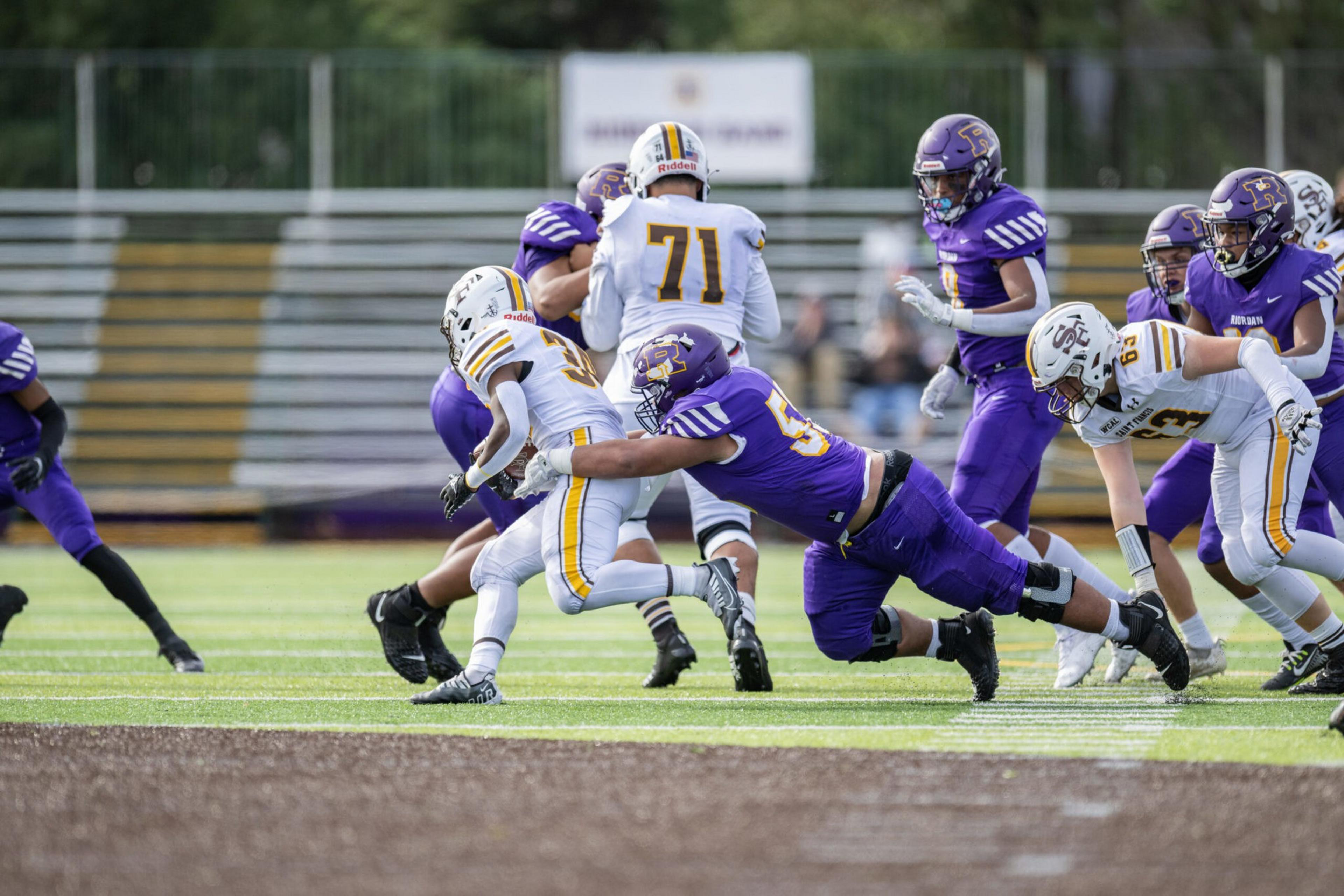 A football player in purple tackles a player in white and yellow as several other players from both teams run nearby on a green field.
