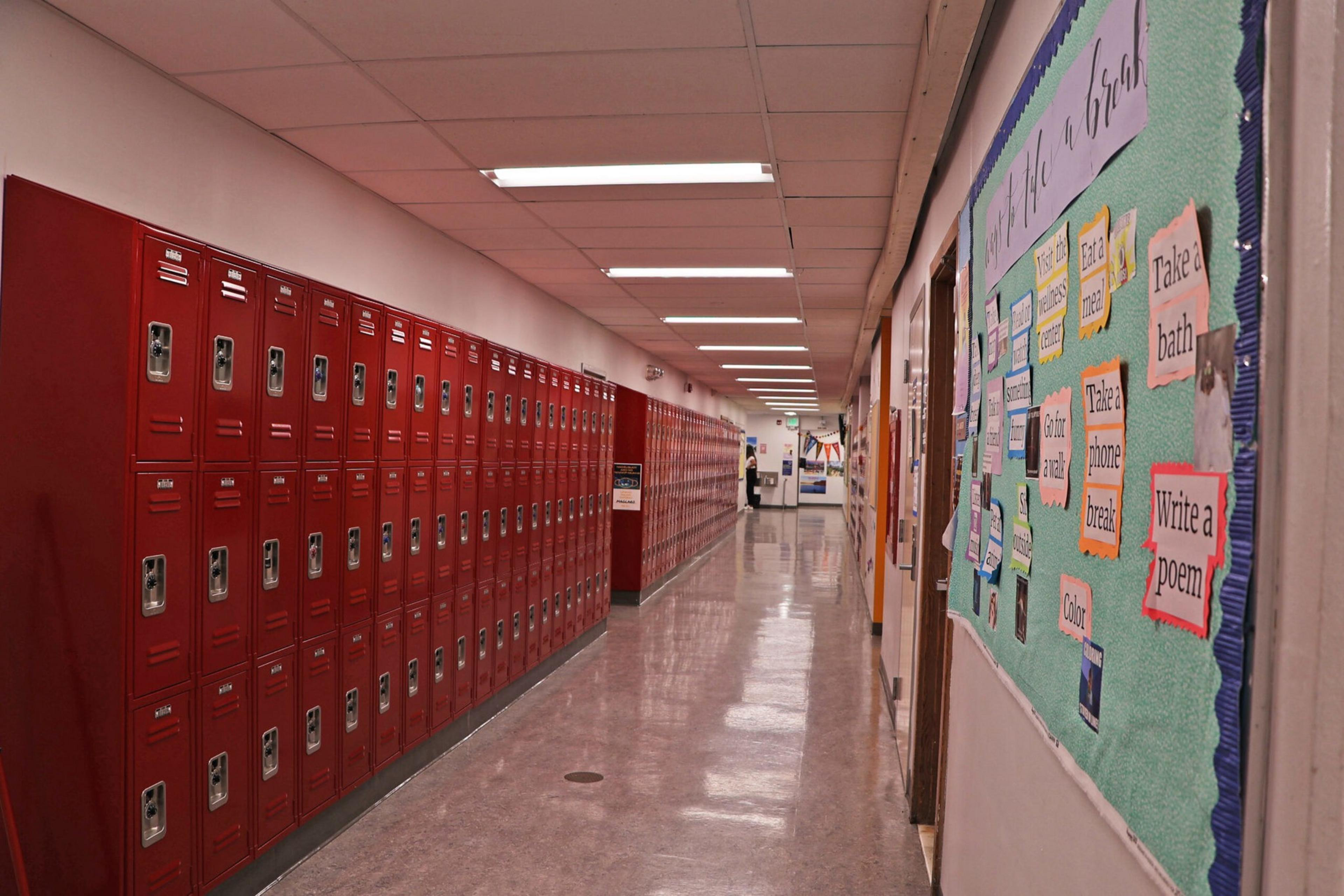 A long hallway with red lockers on the left and a bulletin board with colorful notes on the right, lit by overhead fluorescent lights.