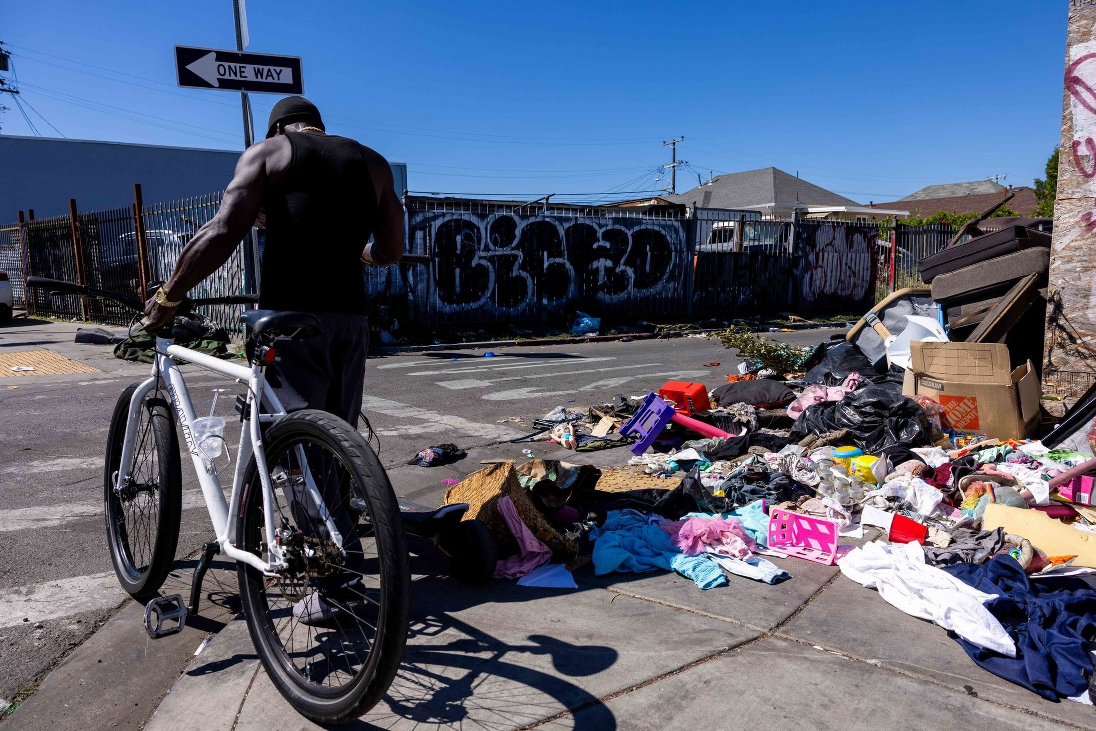 A person with a bicycle stands near a street corner covered with scattered trash, clothes, boxes, and debris under a clear blue sky.