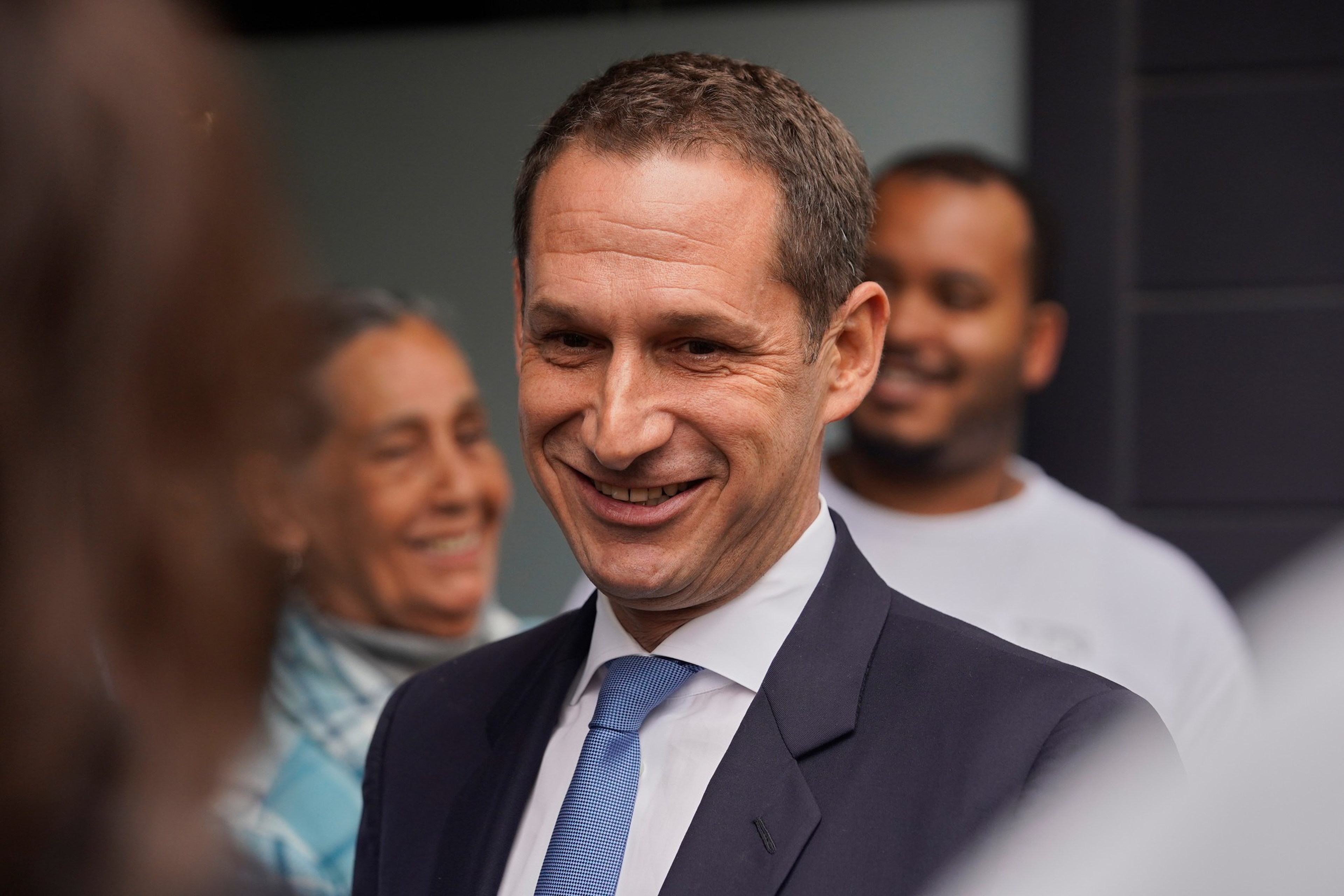 A man in a suit and blue tie smiles warmly, with two blurred people behind him also smiling in a casual setting.