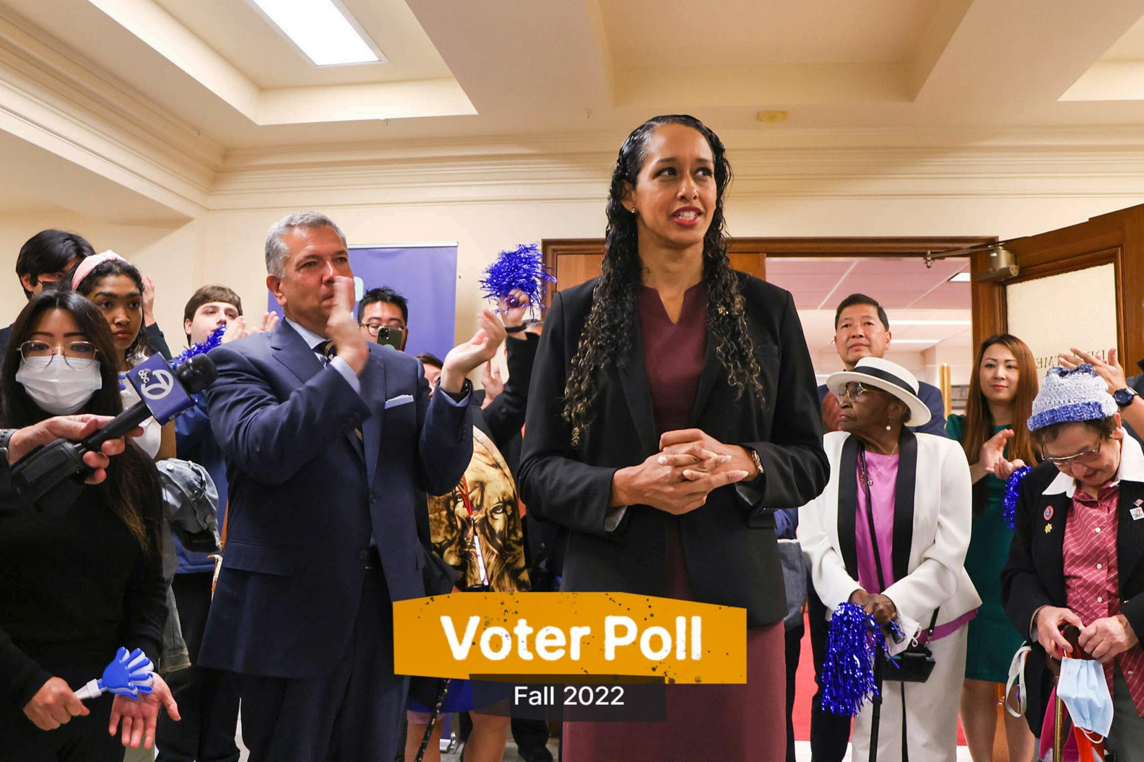 A woman stands speaking in a room full of people applauding and holding blue pom-poms, with a "Voter Poll Fall 2022" sign displayed.