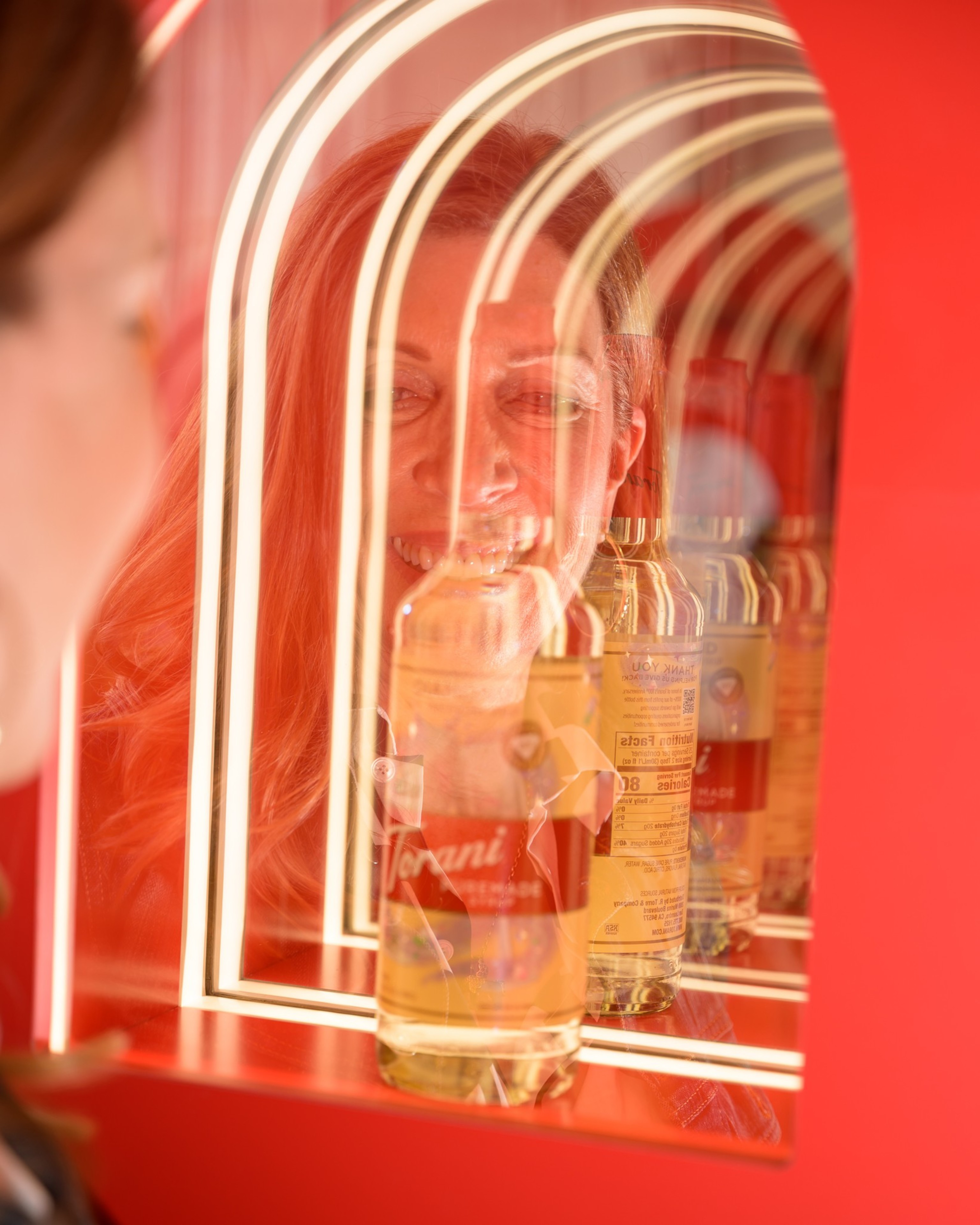 A smiling person is reflected in a neon-lit mirror arch, with Torani syrup bottles in the foreground, creating a colorful, whimsical effect.
