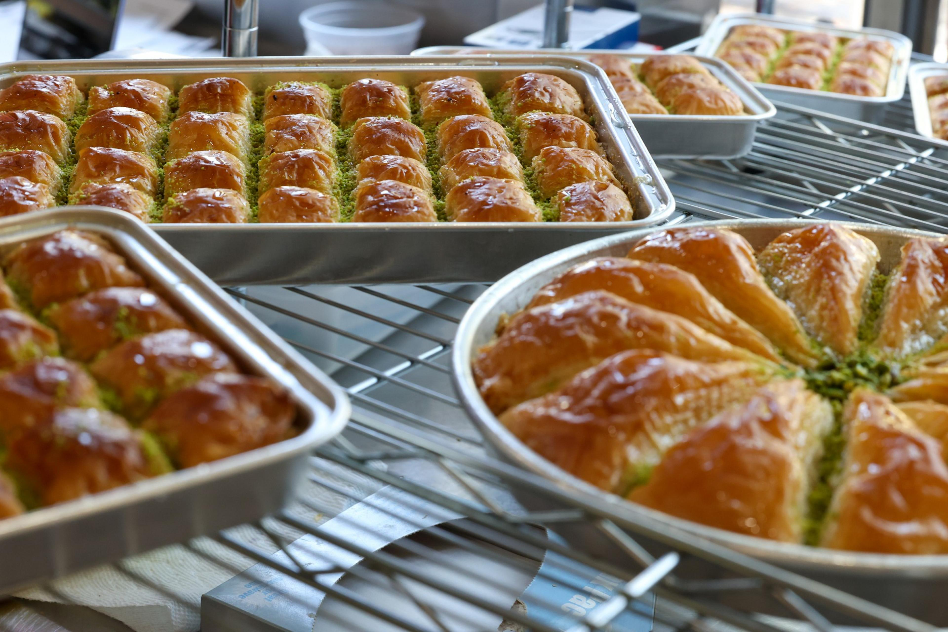 Trays of freshly baked golden-brown baklava with a glossy finish and green pistachio filling, arranged in rectangular and round pans.
