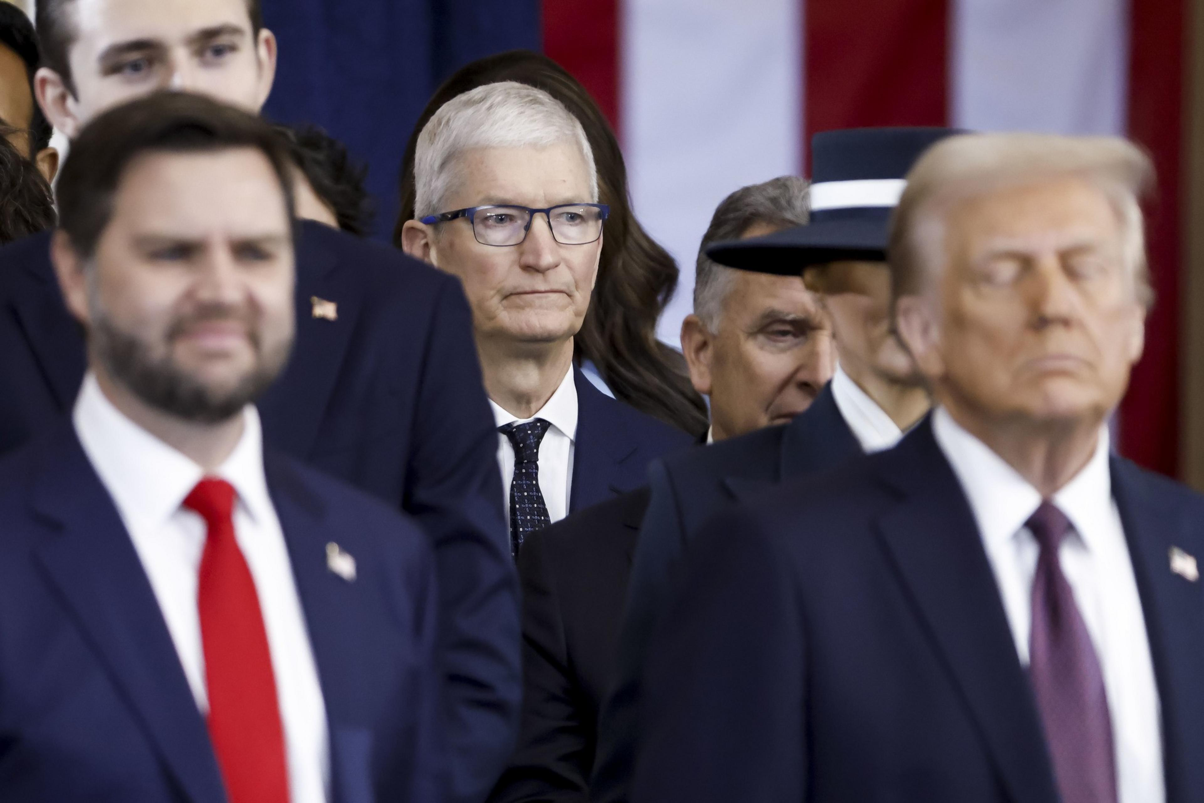 A group of men in suits, some with serious expressions, stand closely together. One wears glasses. A blurred striped background is visible.