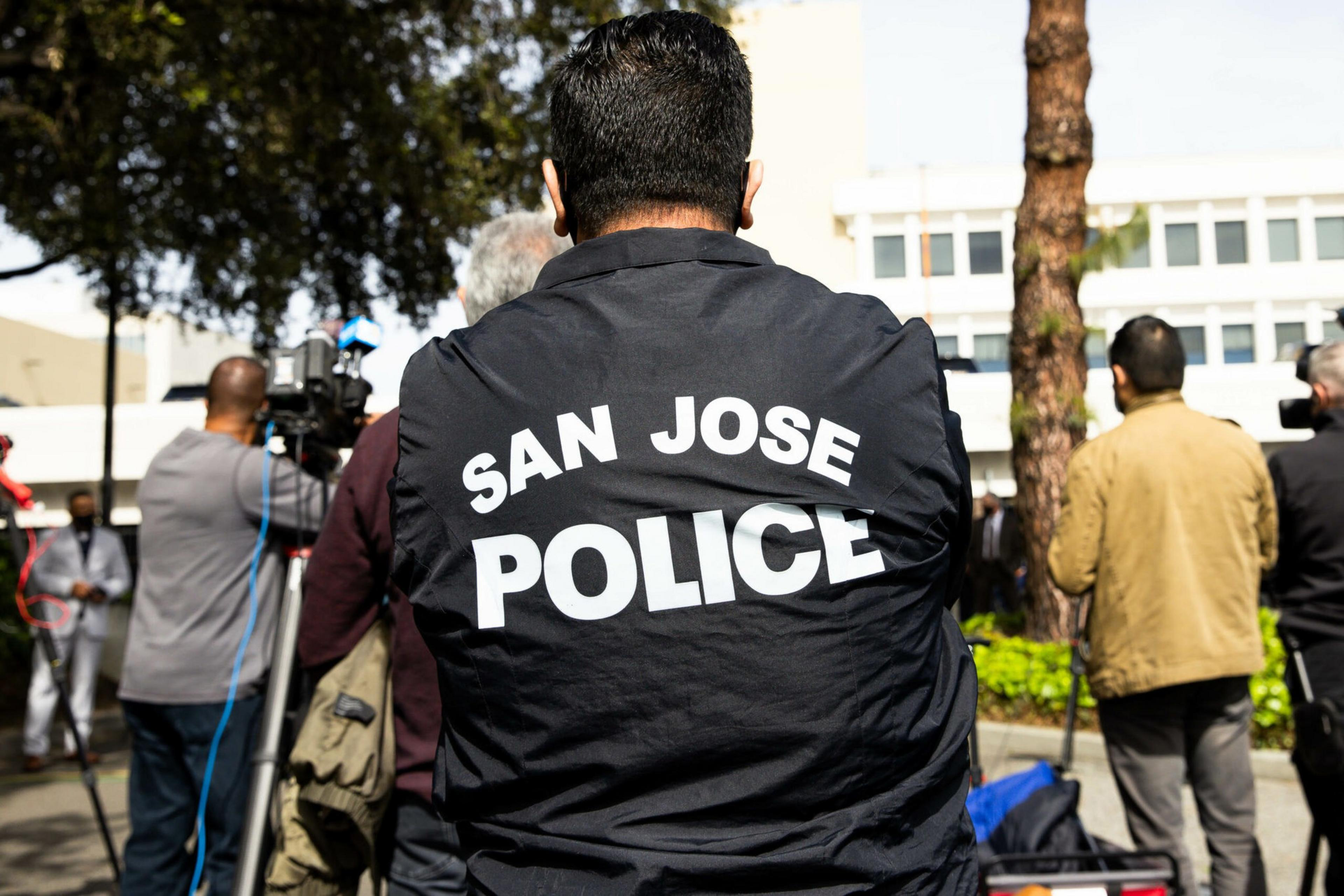 A person wearing a black jacket with "SAN JOSE POLICE" in white letters stands facing a group of people and a camera outdoors.
