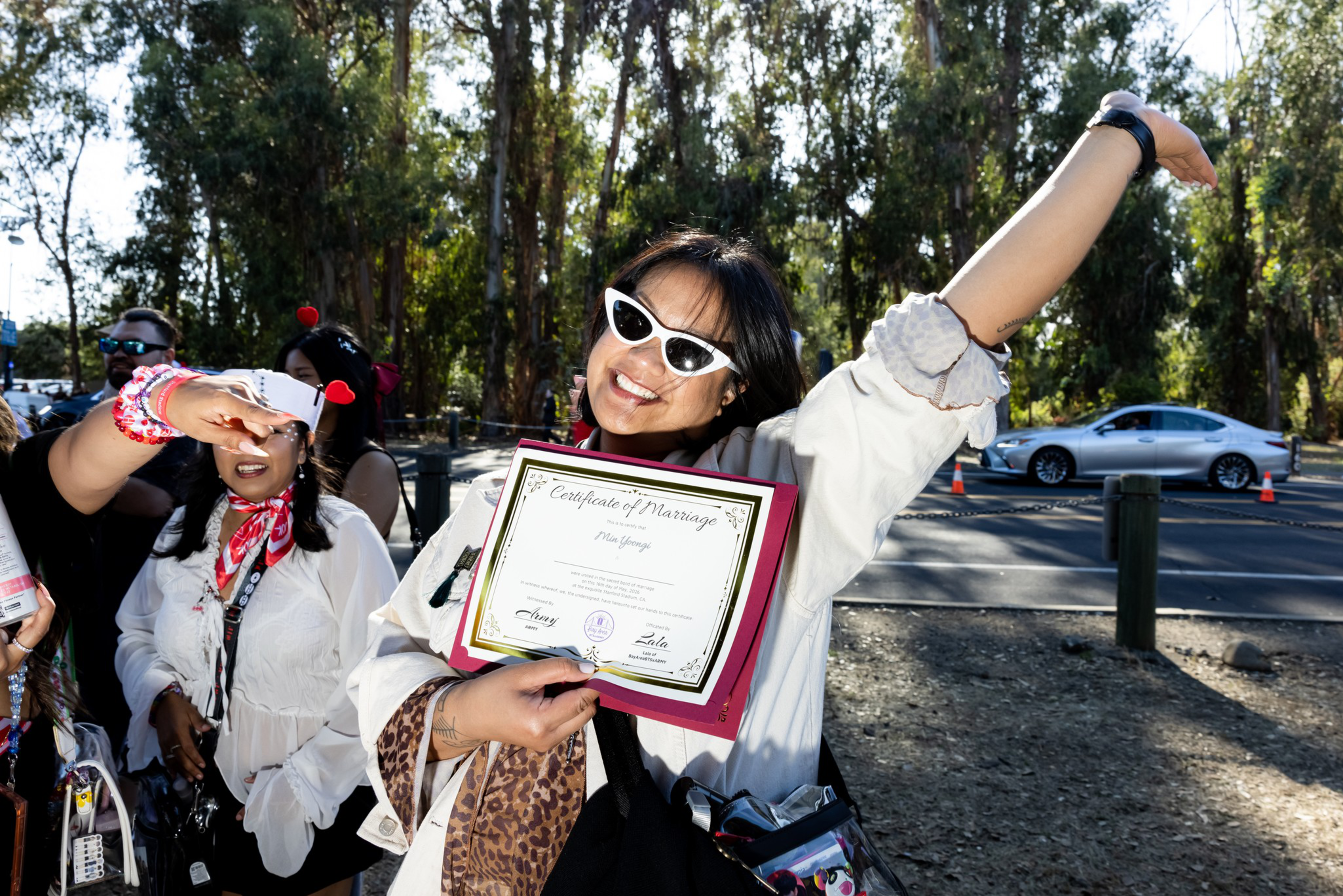 A smiling woman wearing white sunglasses holds up a certificate of marriage, raising one arm in celebration, with trees and a car in the background.