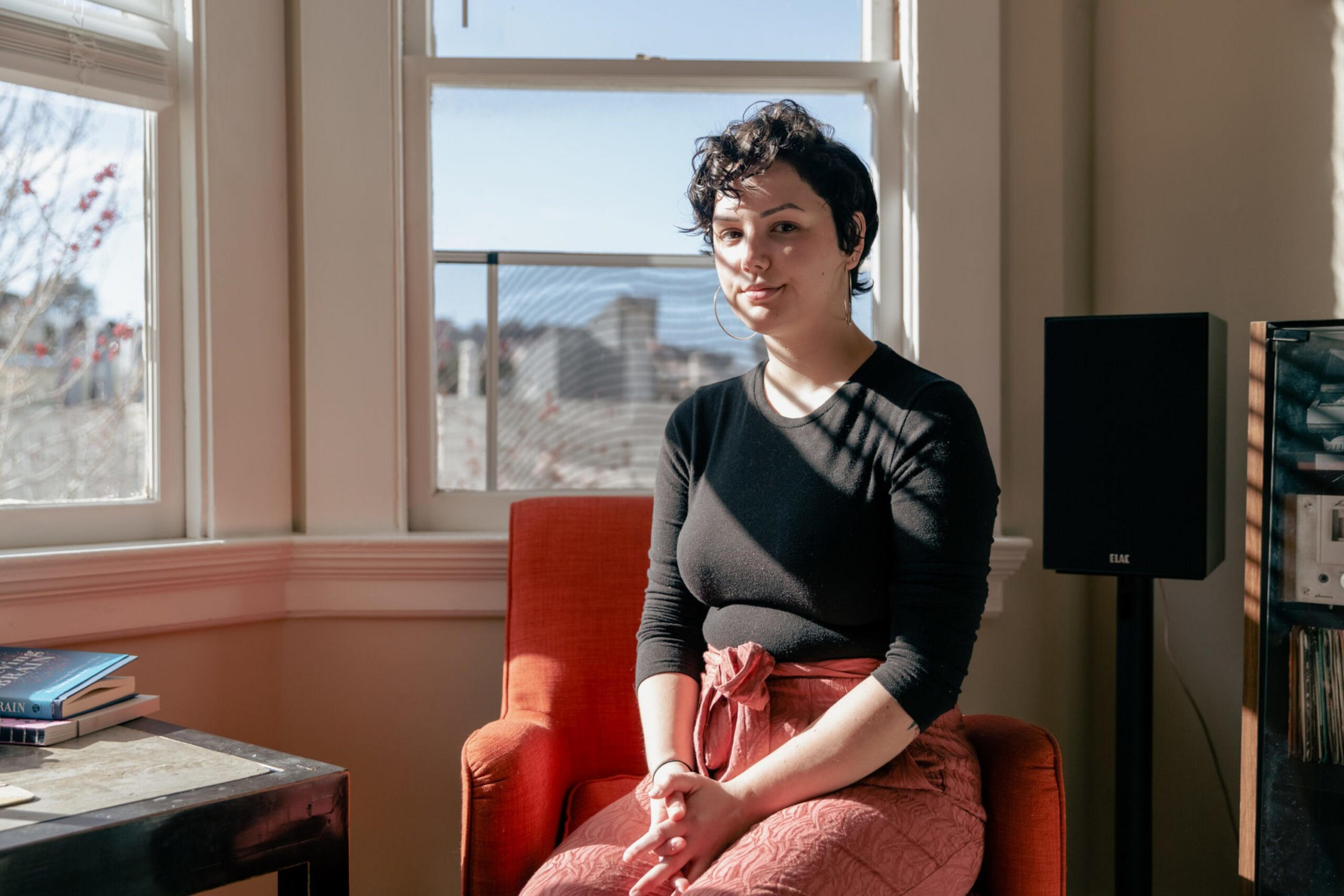 A person with short curly hair sits on an orange chair in a sunlit room with large windows and a speaker nearby.