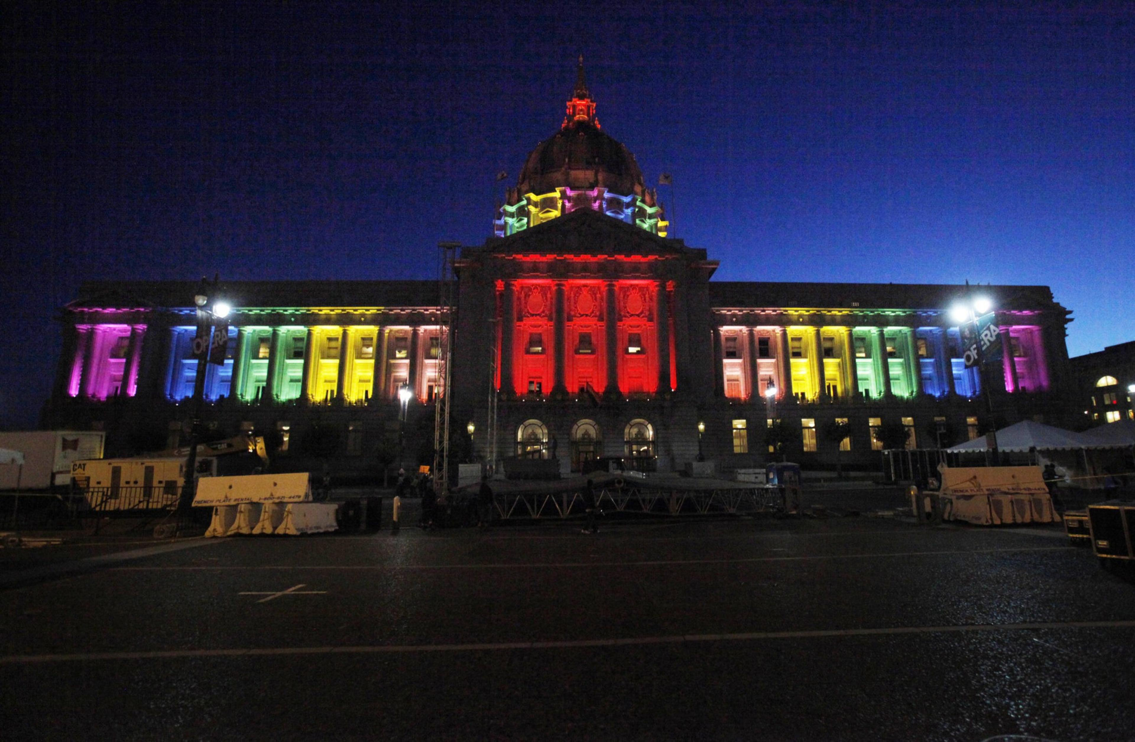 A large classical building is illuminated at night with rainbow colors, transitioning from purple to red and green across its columns and dome.