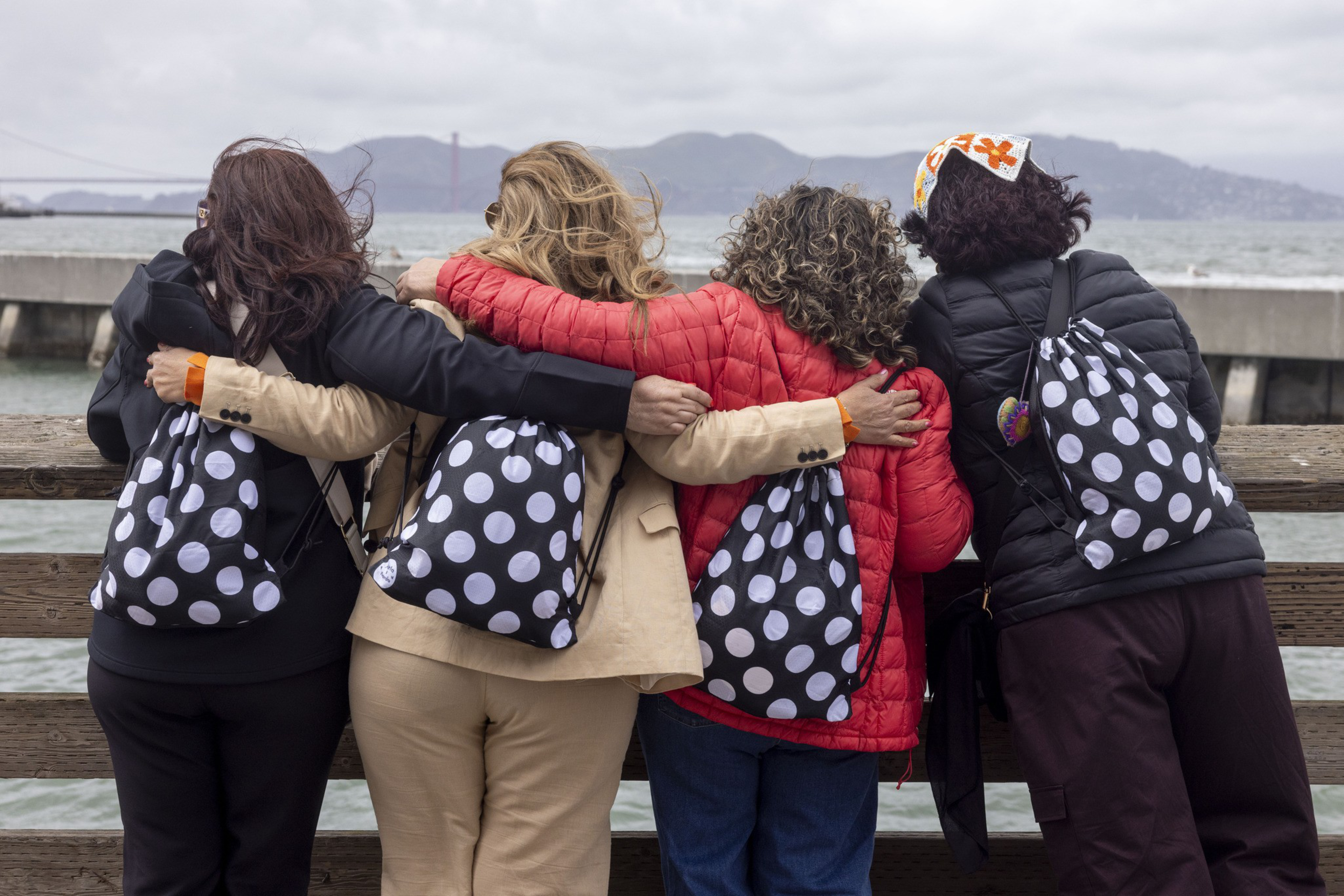 Four people stand arm-in-arm by the water, facing away, each wearing a black and white polka dot bag. They're dressed warmly, with jackets and hats.