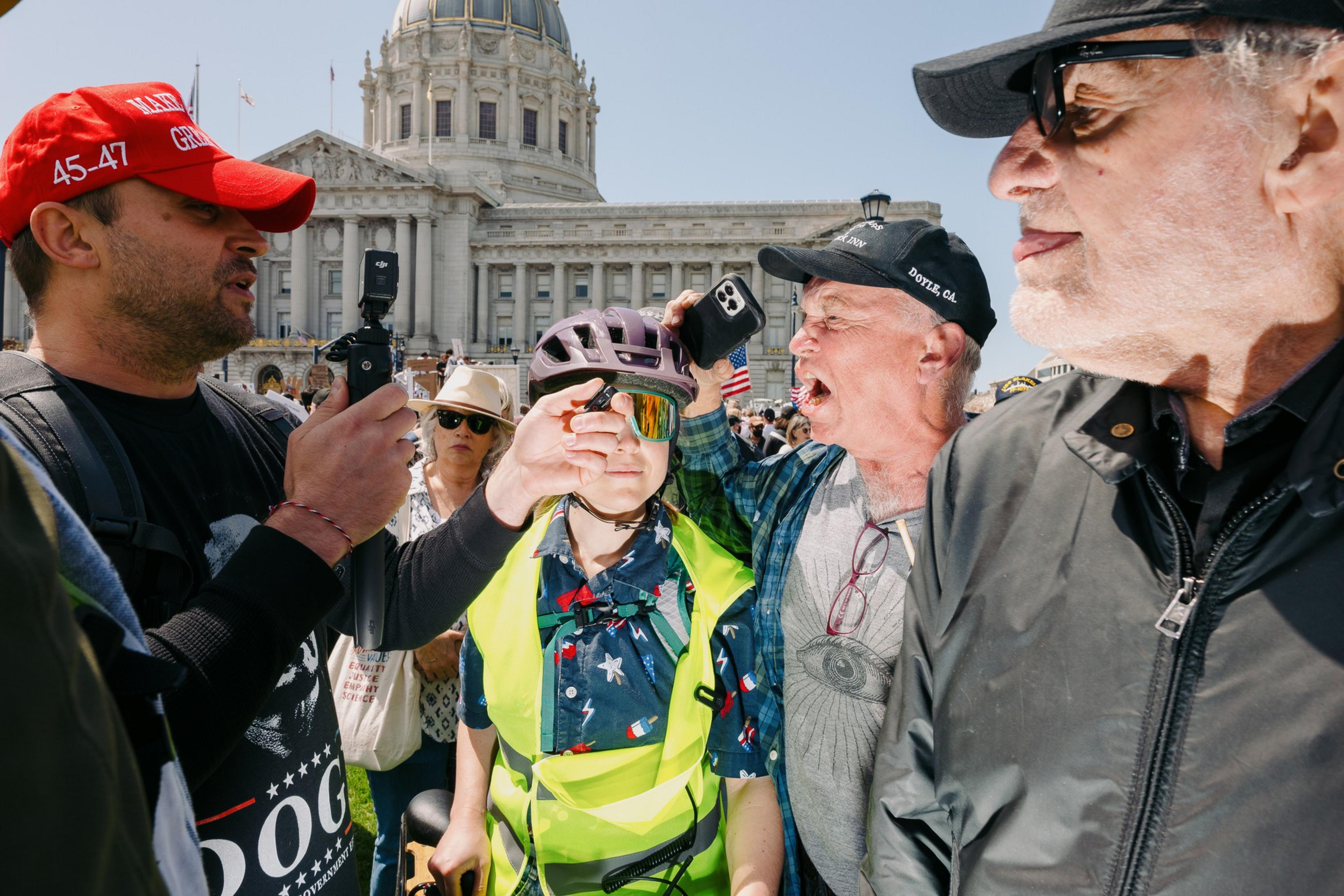 A group of people are engaged in a heated discussion outside a large, domed building. One person wears a red cap, another holds a phone, and a young person in a helmet stands in the middle.