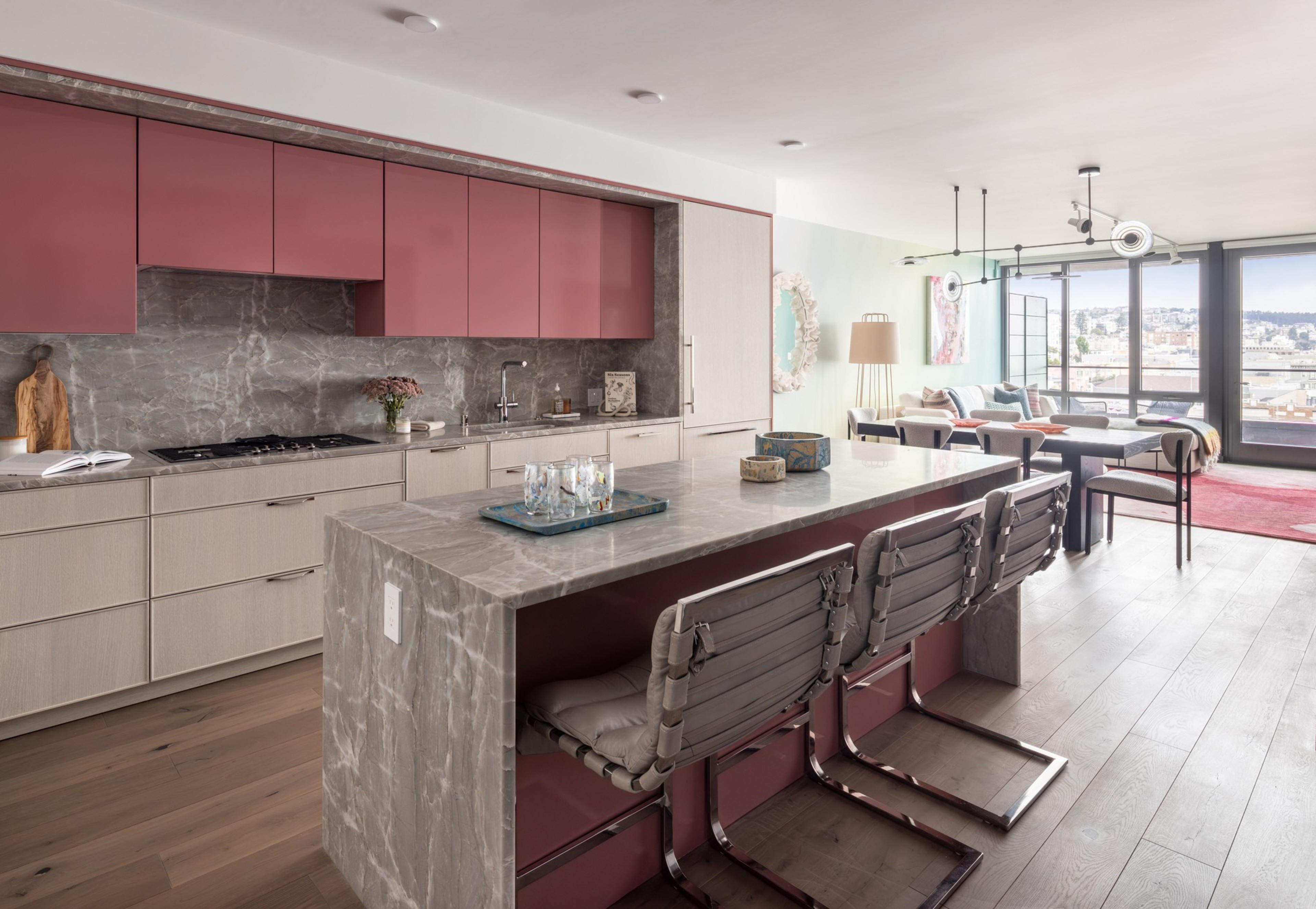 A modern kitchen with pink upper cabinets, light wood lower cabinets, a gray marble island with cushioned chairs, and a bright living area with large windows beyond.