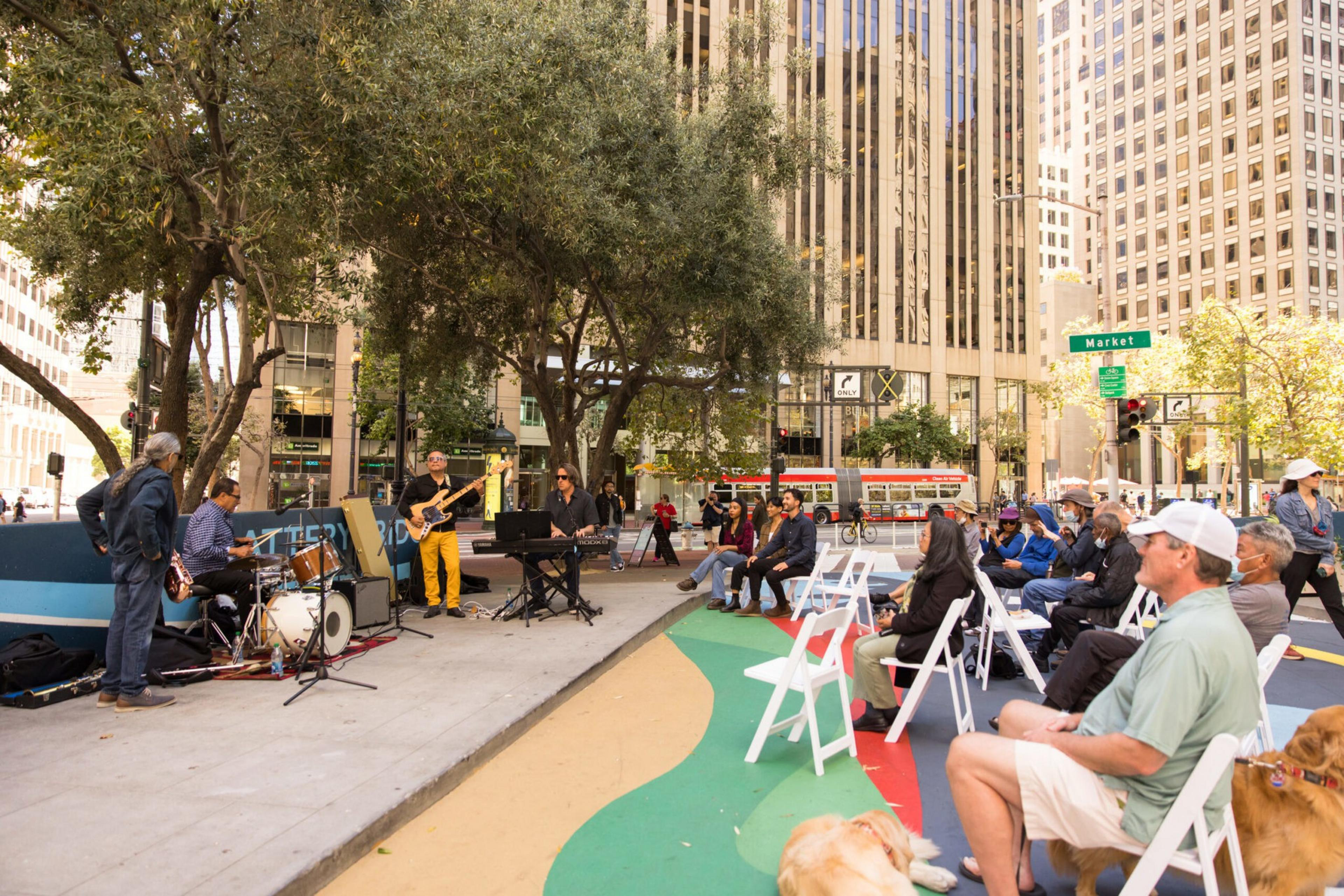 A small band plays jazz on a city sidewalk while an audience sits on white chairs under trees among tall buildings.