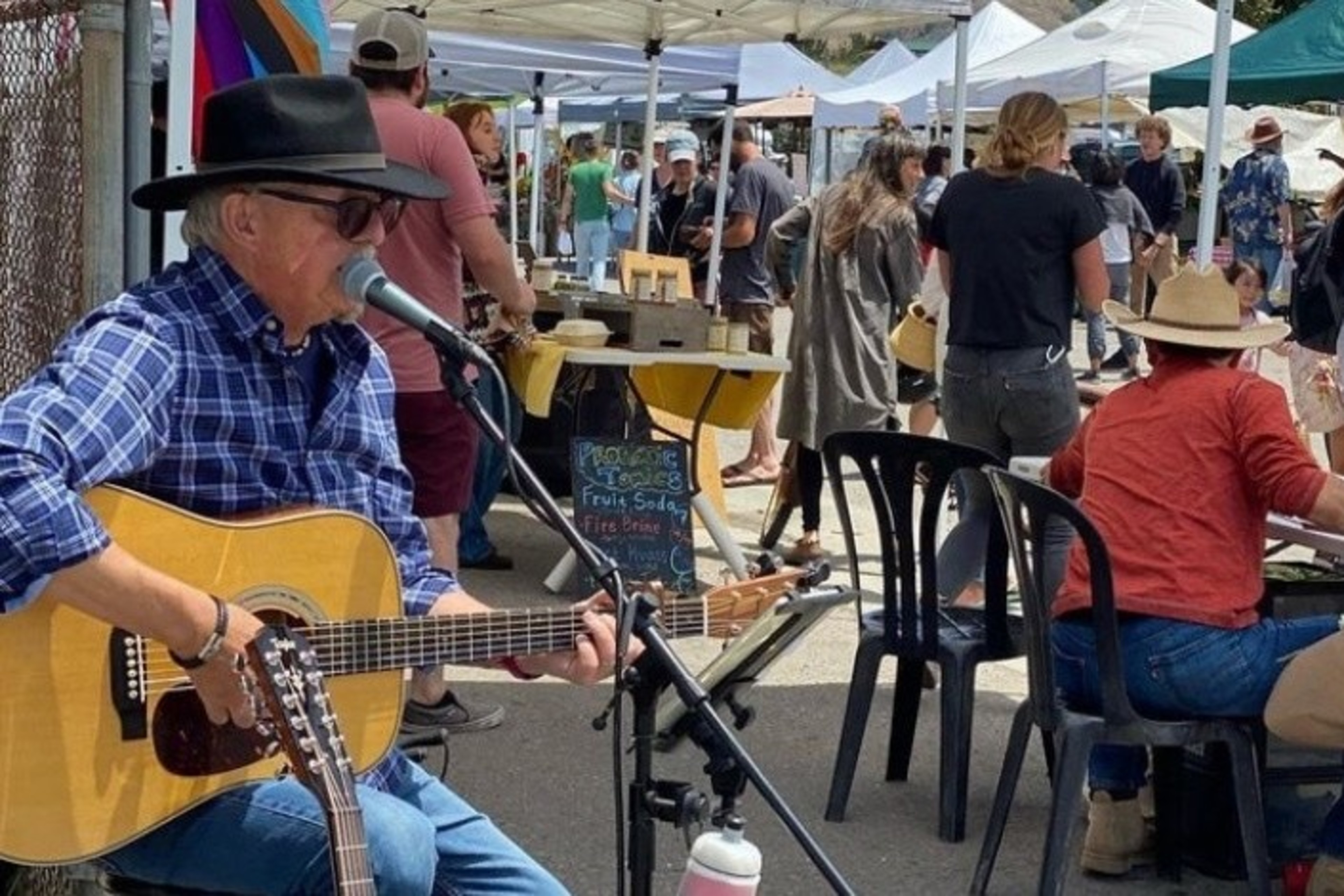 A guitar player sings into a microphone at an outdoor market, while people walk by tents. In the foreground, a person in a hat sits at a table.