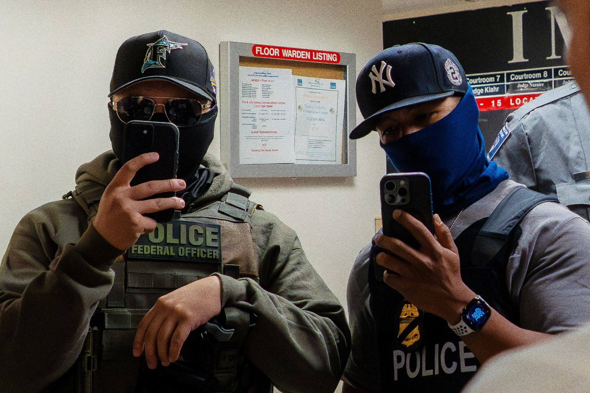 Two masked men wearing police vests, hats, and sunglasses take a mirror selfie with smartphones in a hallway labeled "Floor Warden Listing."