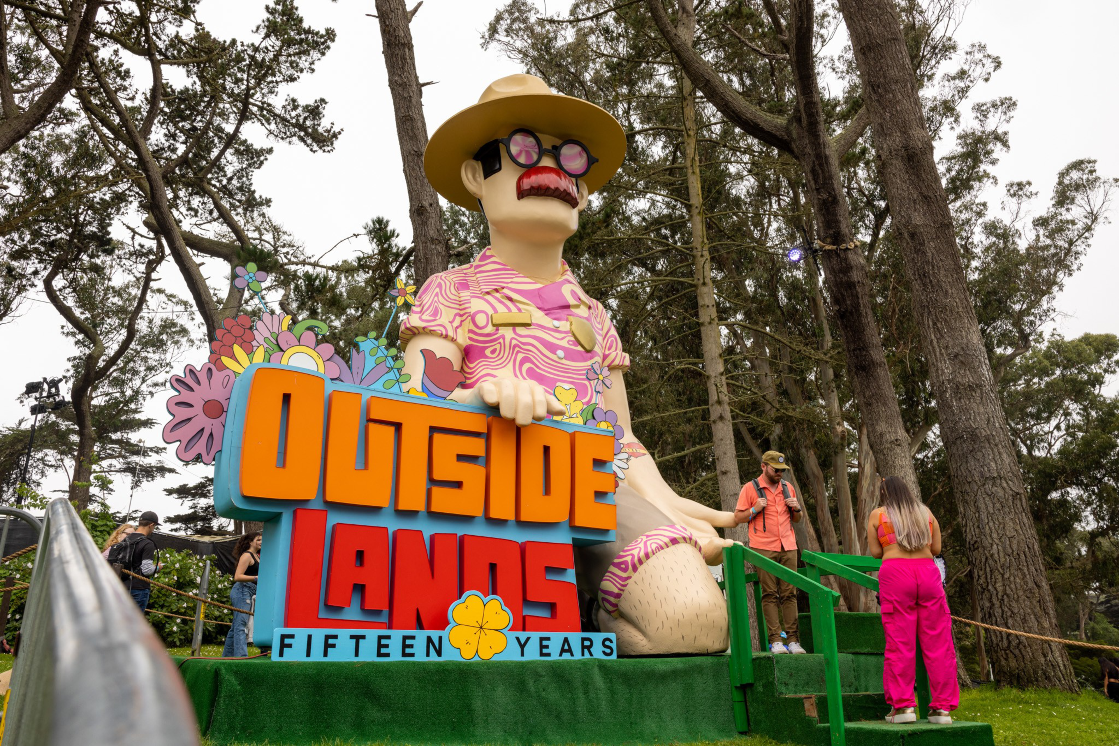 A large colorful statue of a man with pink sunglasses, a mustache, and a hat sits behind a sign that reads "Outside Lands Fifteen Years" among tall trees.