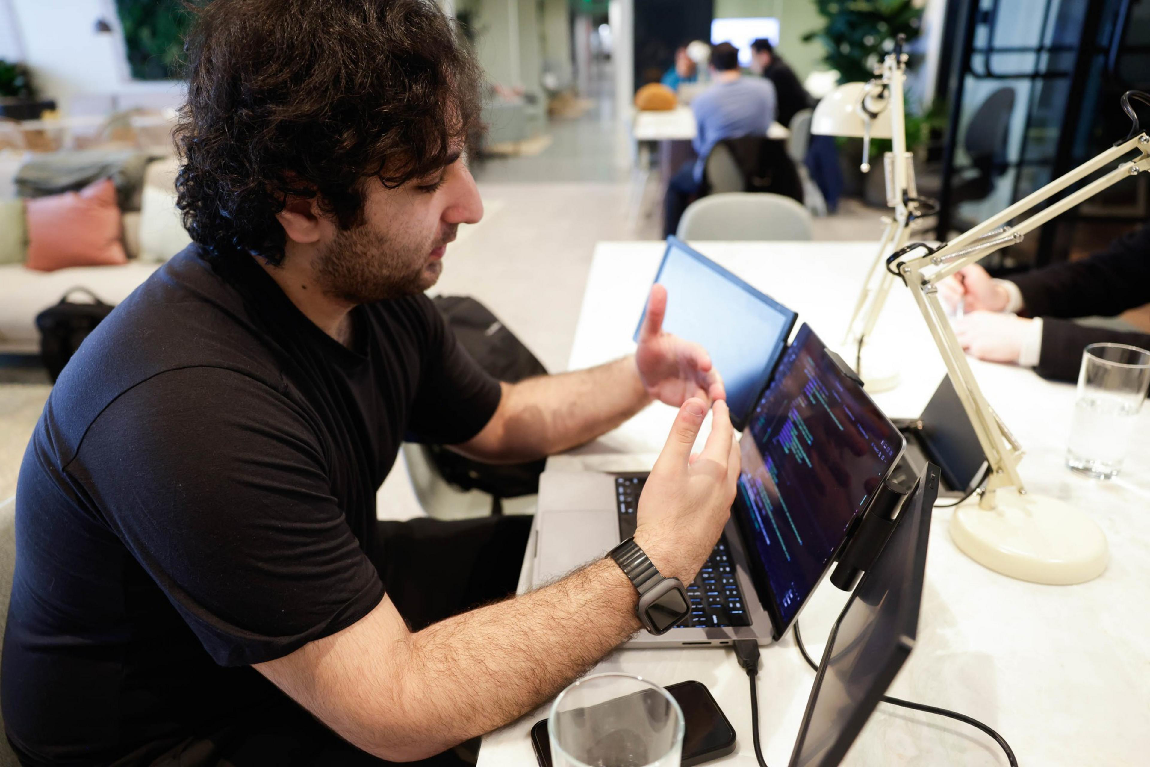 A man gestures at a laptop screen with code, indoors, at a desk with a lamp and glass.